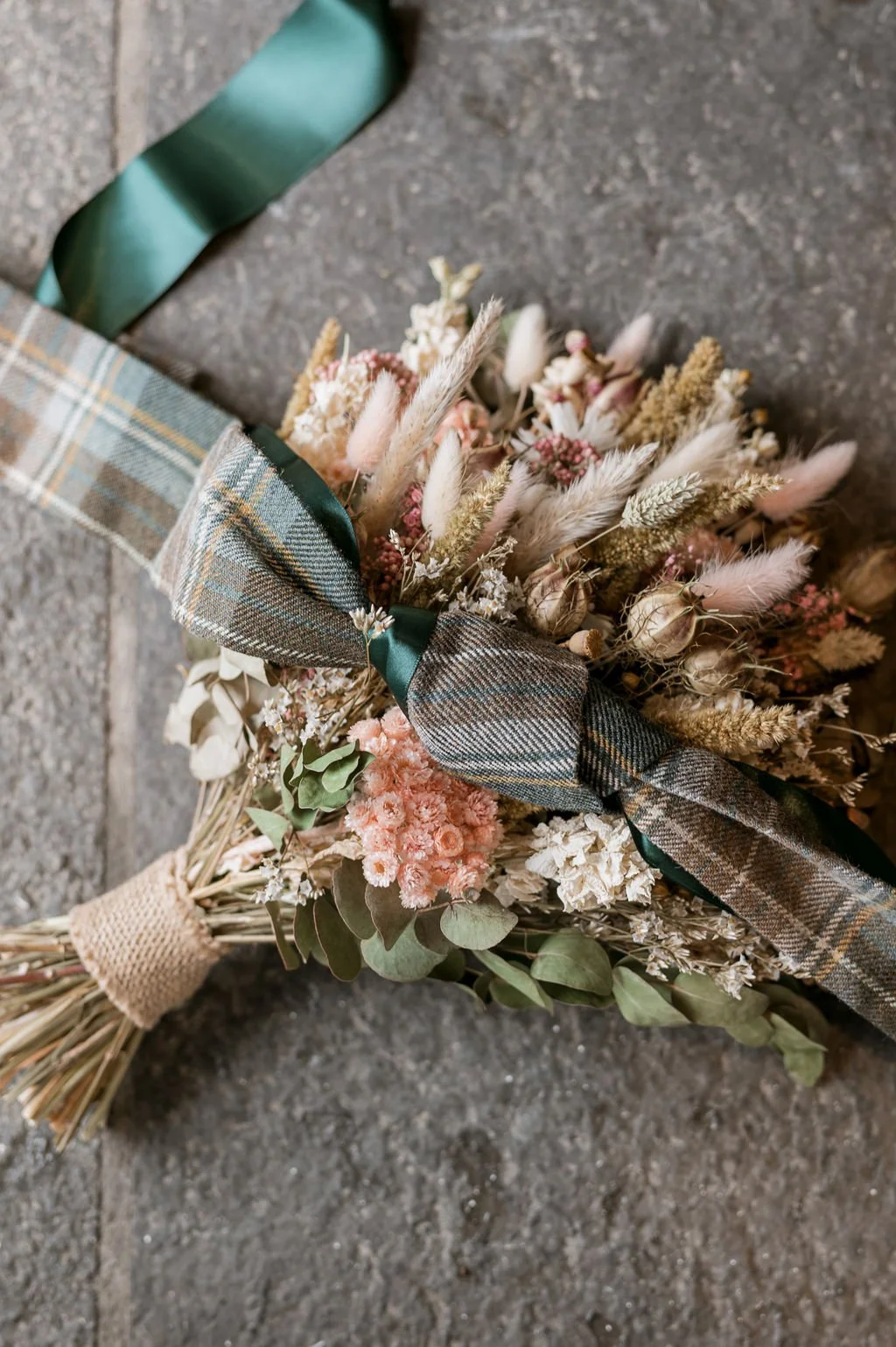 A bouquet of dried flowers with a plaid ribbon and green handle, lying on a stone surface.