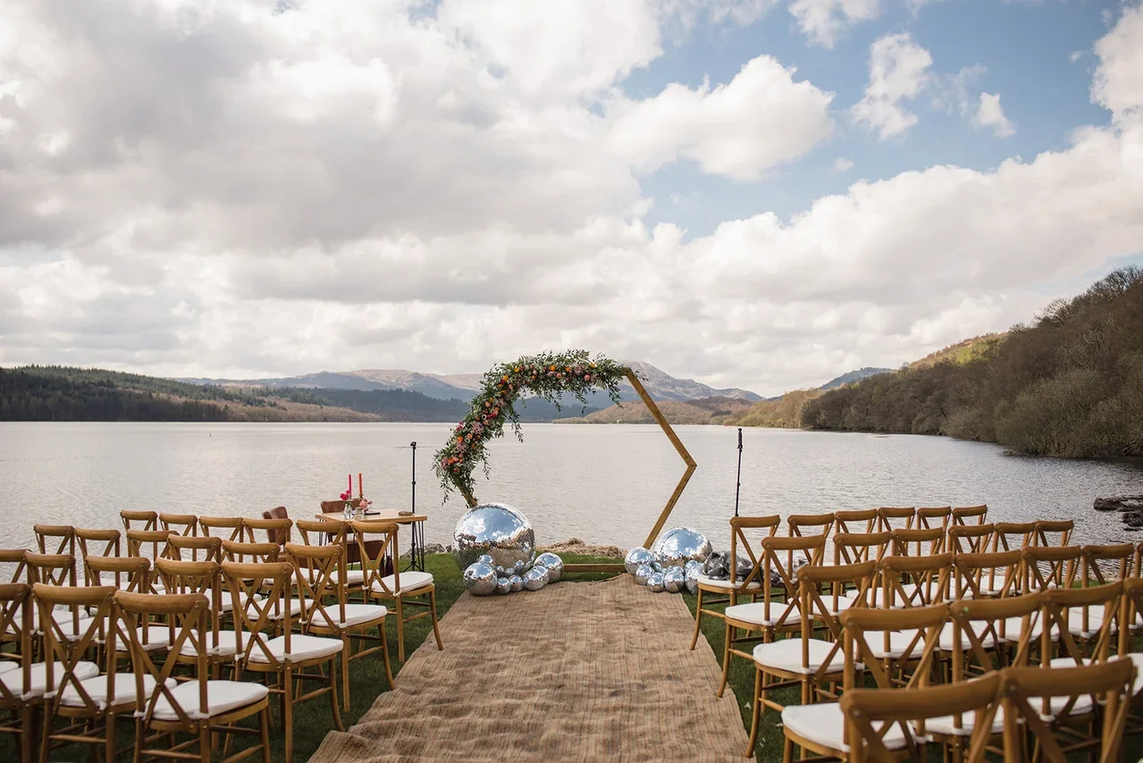 Outdoor wedding setup by a lake with wooden chairs, a flower-decorated hexagon arch, metallic balloons, and scenic mountains in the background.