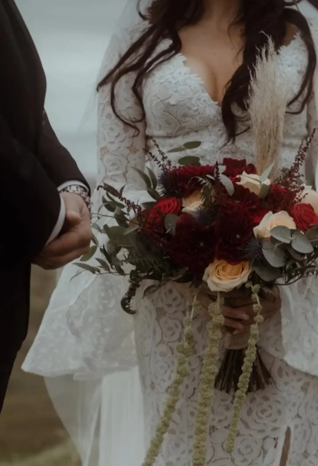 A bride in a white lace wedding dress holding a bouquet of red, cream, and dark purple flowers with green foliage, standing next to a person in a black suit.