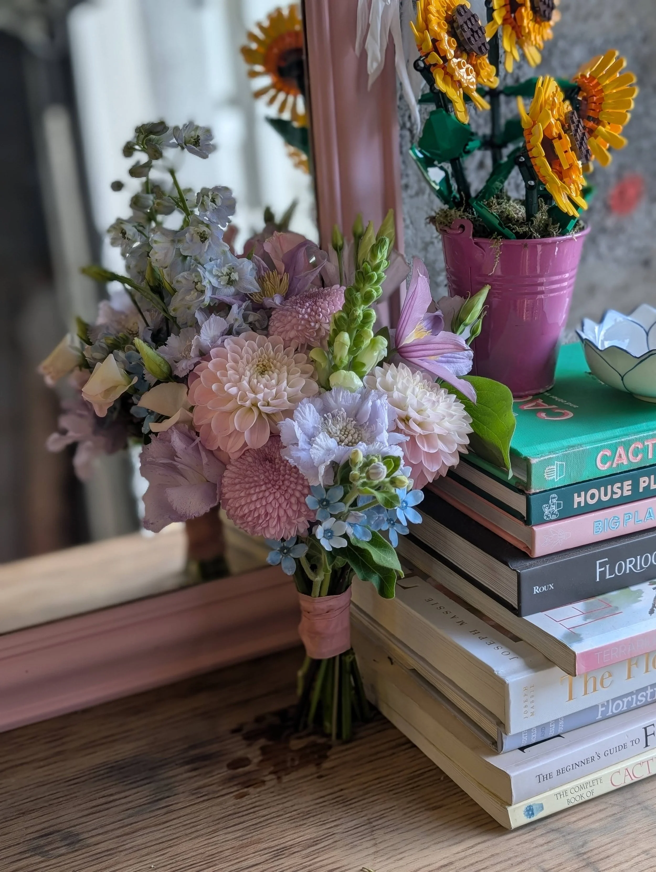 A bouquet of pink, white, and purple flowers sits on a wooden table, with a mirror, pink pot with sunflowers, and a stack of gardening books in the background.