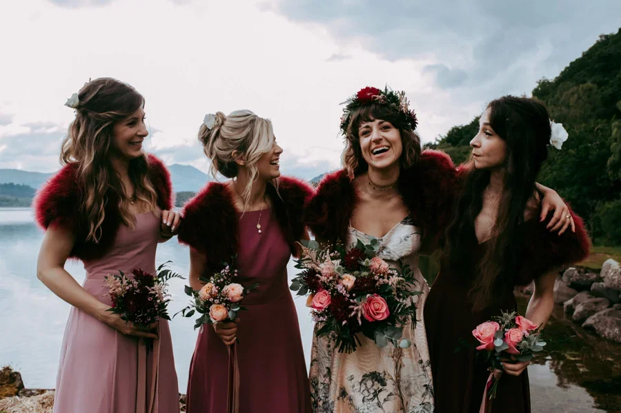 Four women in dresses holding bouquets, standing outdoors near a body of water, celebrating a wedding or special occasion.