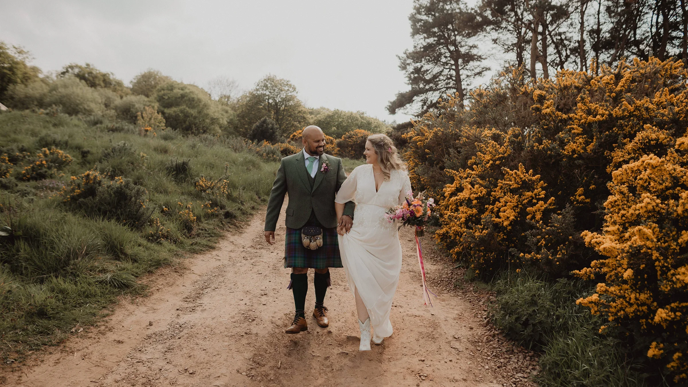 A couple walking hand in hand on a dirt path surrounded by green shrubbery and yellow flowers, the man wearing a kilt and blazer, and the woman in a white dress carrying a bouquet.