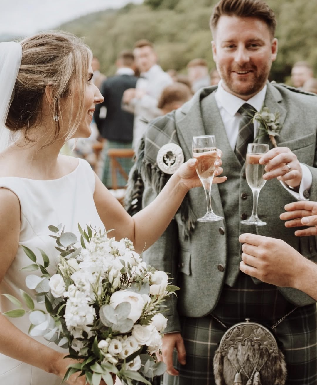 Bride and groom at a wedding celebration holding glasses of champagne, smiling, with guests in the background. The bride is holding a bouquet of white flowers, and the groom is wearing traditional Scottish attire with a kilt.