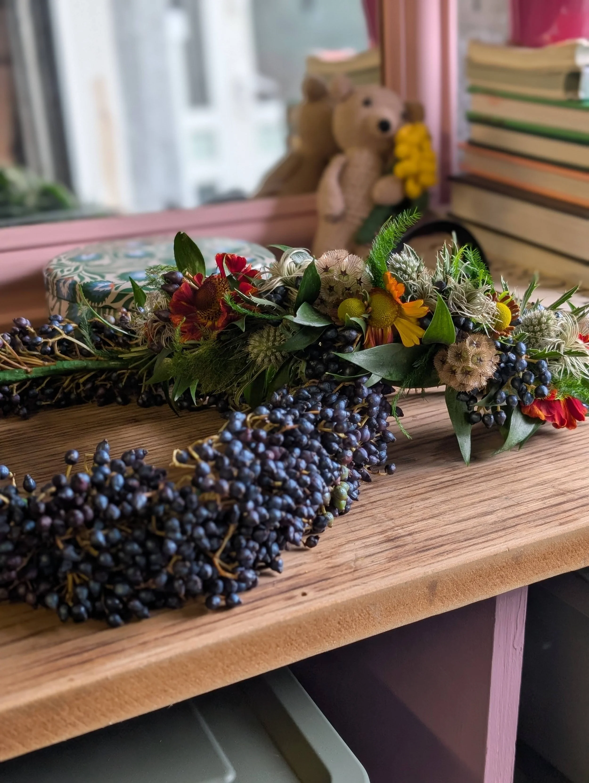 A wooden surface with a floral crown made of berries, leaves, and flowers. In the background, a mirror reflecting two teddy bears, books, and a window.