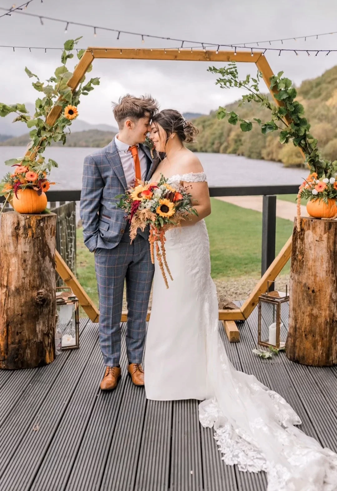 A bride and groom standing close together outdoors by a lake during their wedding, with a wooden arch decorated with sunflowers and greenery behind them, and lanterns on tree stumps on either side.