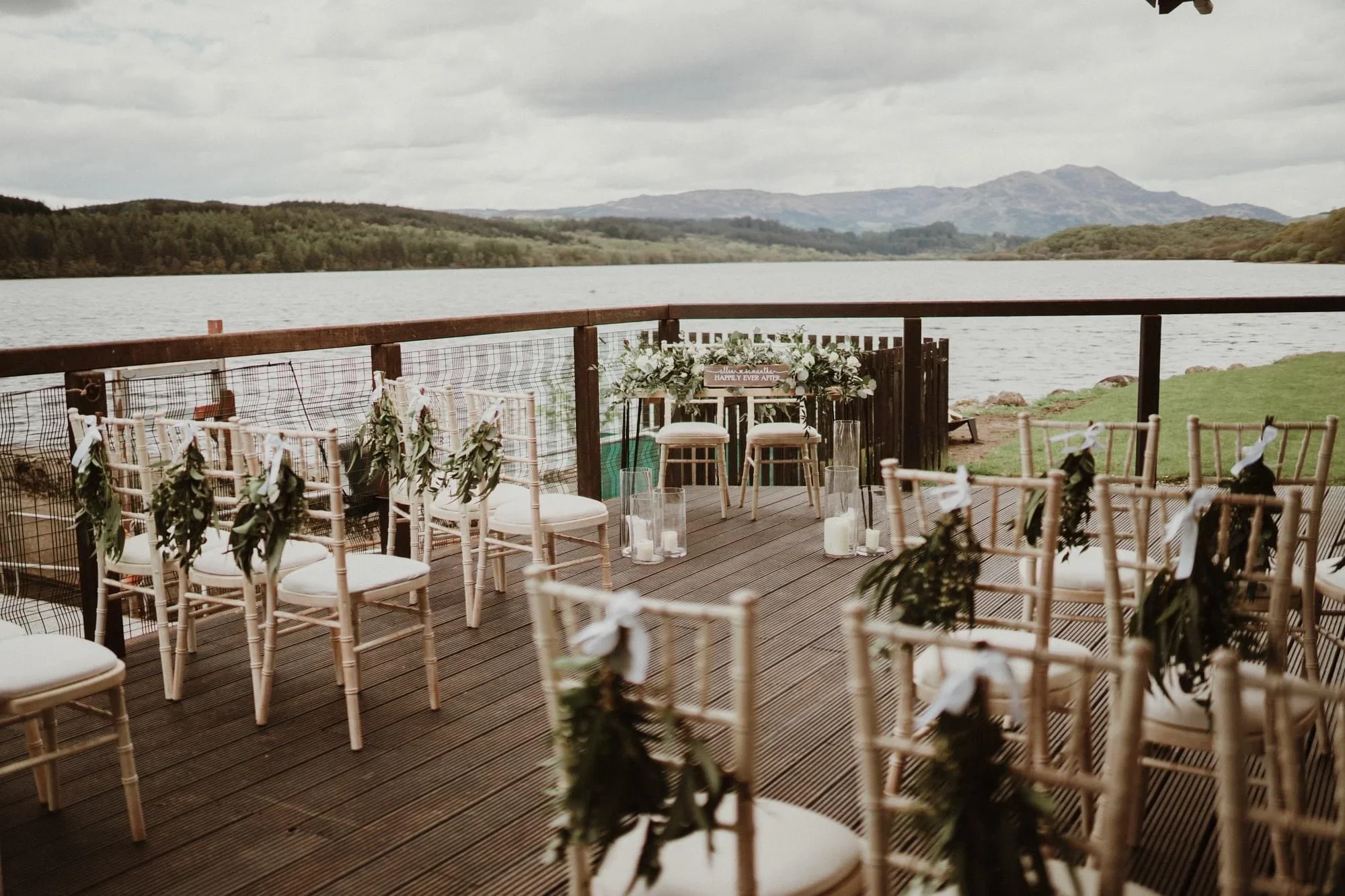 Outdoor wedding setup on a deck overlooking a lake with chairs decorated with greenery and white ribbons, and a floral arch in the background with a lake and hills in the distance.