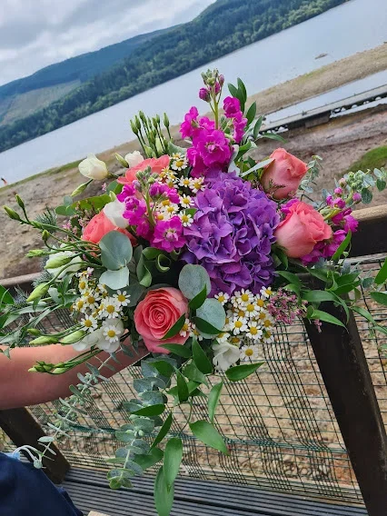 Colorful bouquet of pink roses, purple hydrangea, white daisies, and other greenery held outdoors near a lake with mountains in the background.