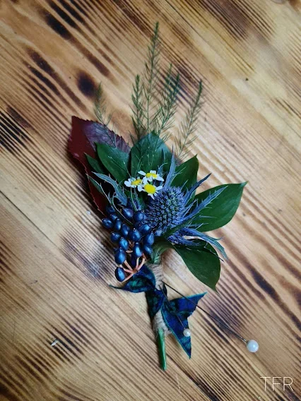 A boutonniere with green leaves, blue thistle, small white and yellow flowers, and blue berries, pinned on a wooden surface.