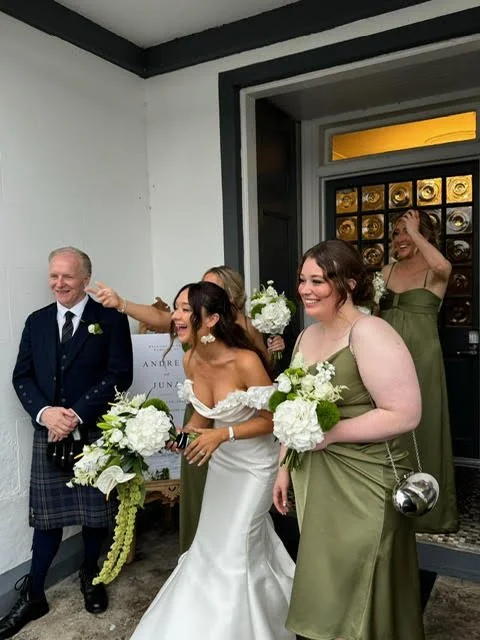 Bride in white wedding gown surrounded by bridesmaids in green dresses, all holding bouquets of white flowers, inside a building with a black and white interior.