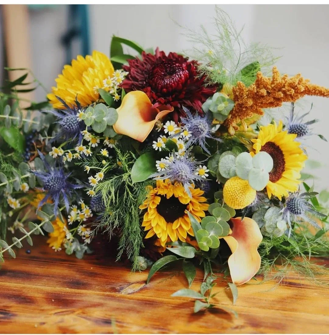 A colorful bouquet of flowers featuring sunflowers, calla lilies, dahlias, and various greenery on a wooden surface.