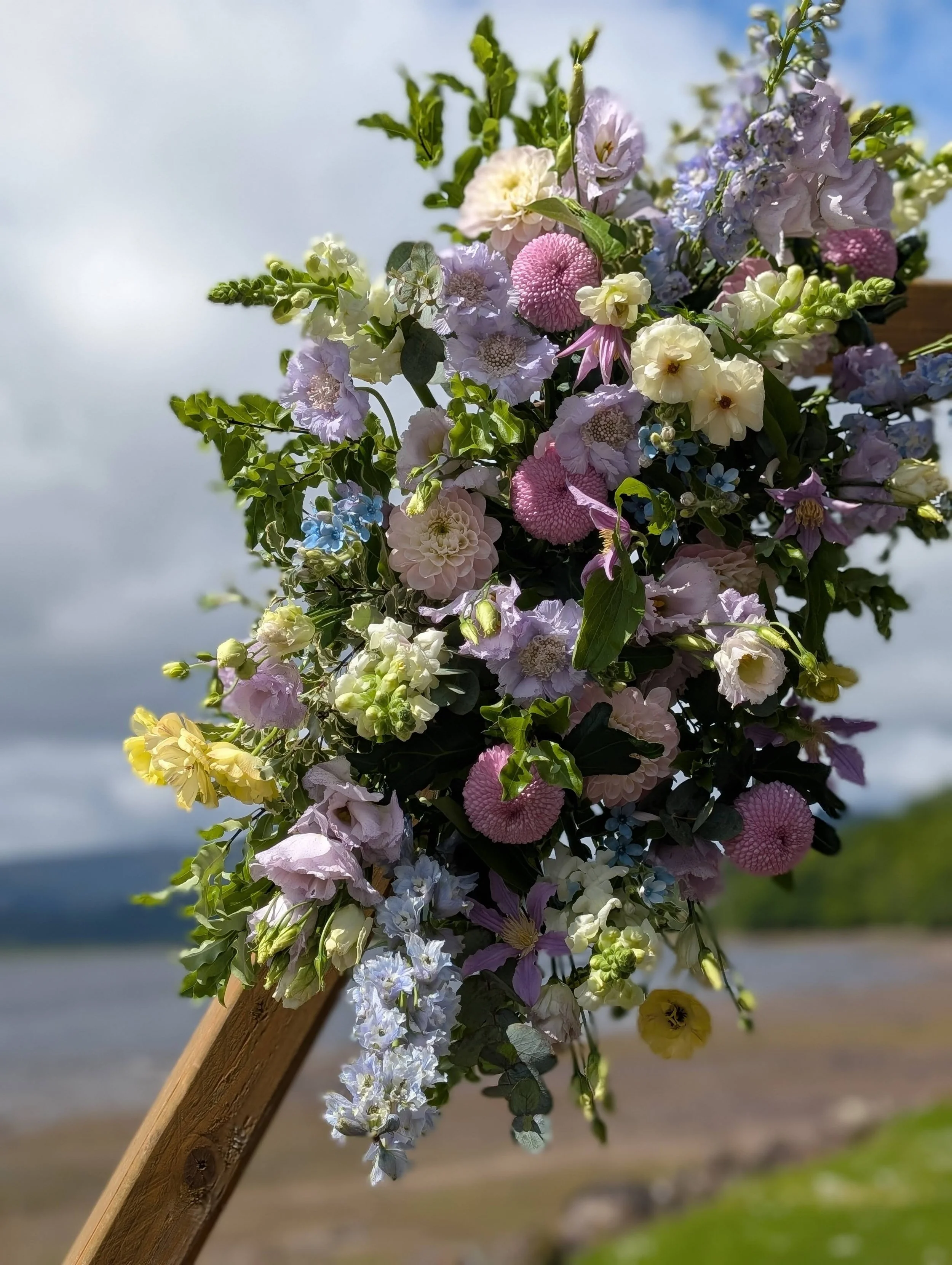 A bouquet of colorful flowers, including pink, yellow, blue, and white blossoms, attached to a wooden stick with a natural outdoor background of sky, water, and trees.
