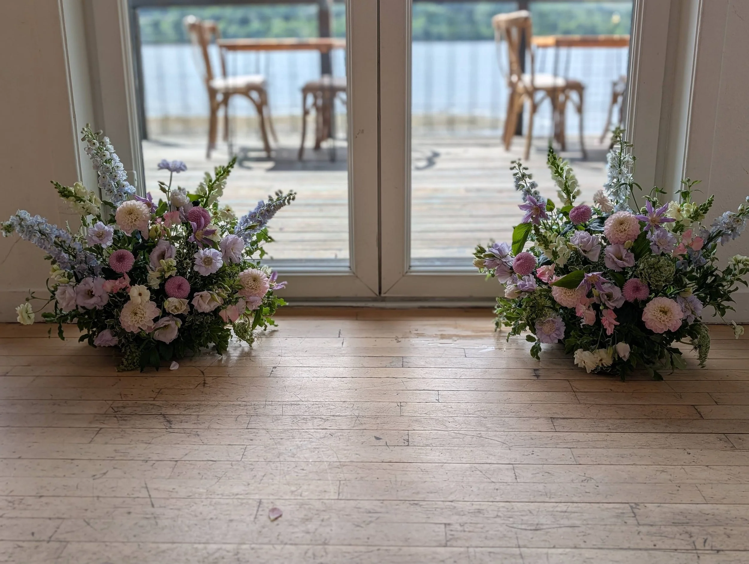 Two floral arrangements with pink, purple, and white flowers sitting on a wooden floor near a glass door leading to an outdoor patio with chairs and a railing overlooking water.