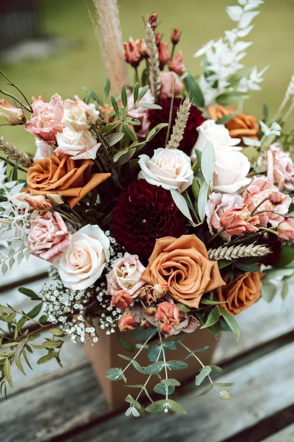 Close-up of a bouquet of mixed flowers including roses, dahlias, and other blooms in orange, white, pink, and burgundy, with greenery and dried elements. outdoor setting with grass background.