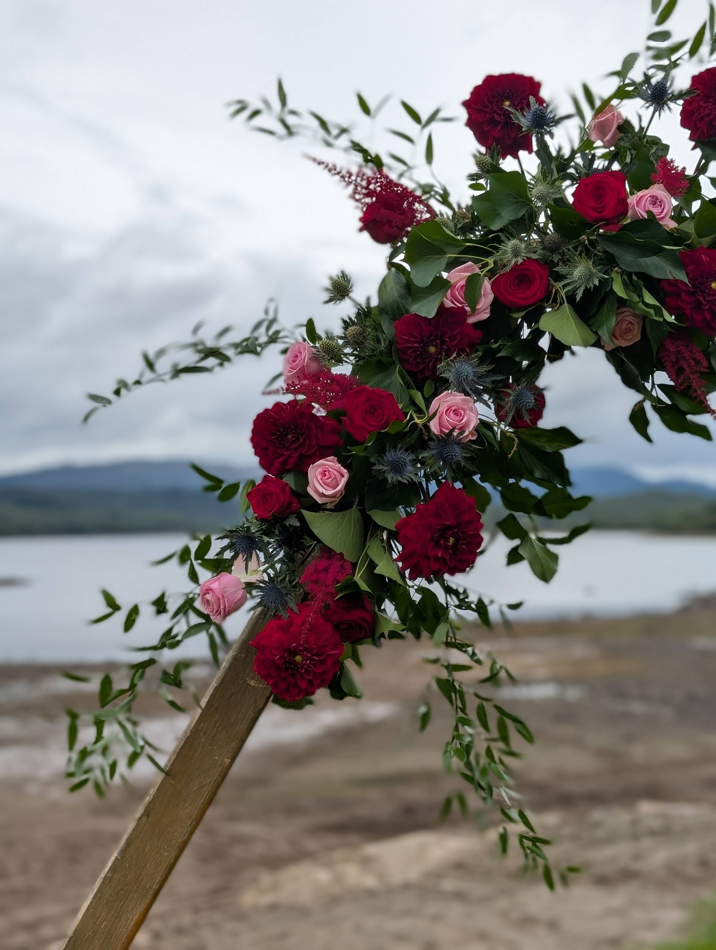 A floral arrangement with red and pink roses, dark red dahlias, blue thistle flowers, and green leaves, held on a wooden stick outdoors near water with mountains and a cloudy sky in the background.