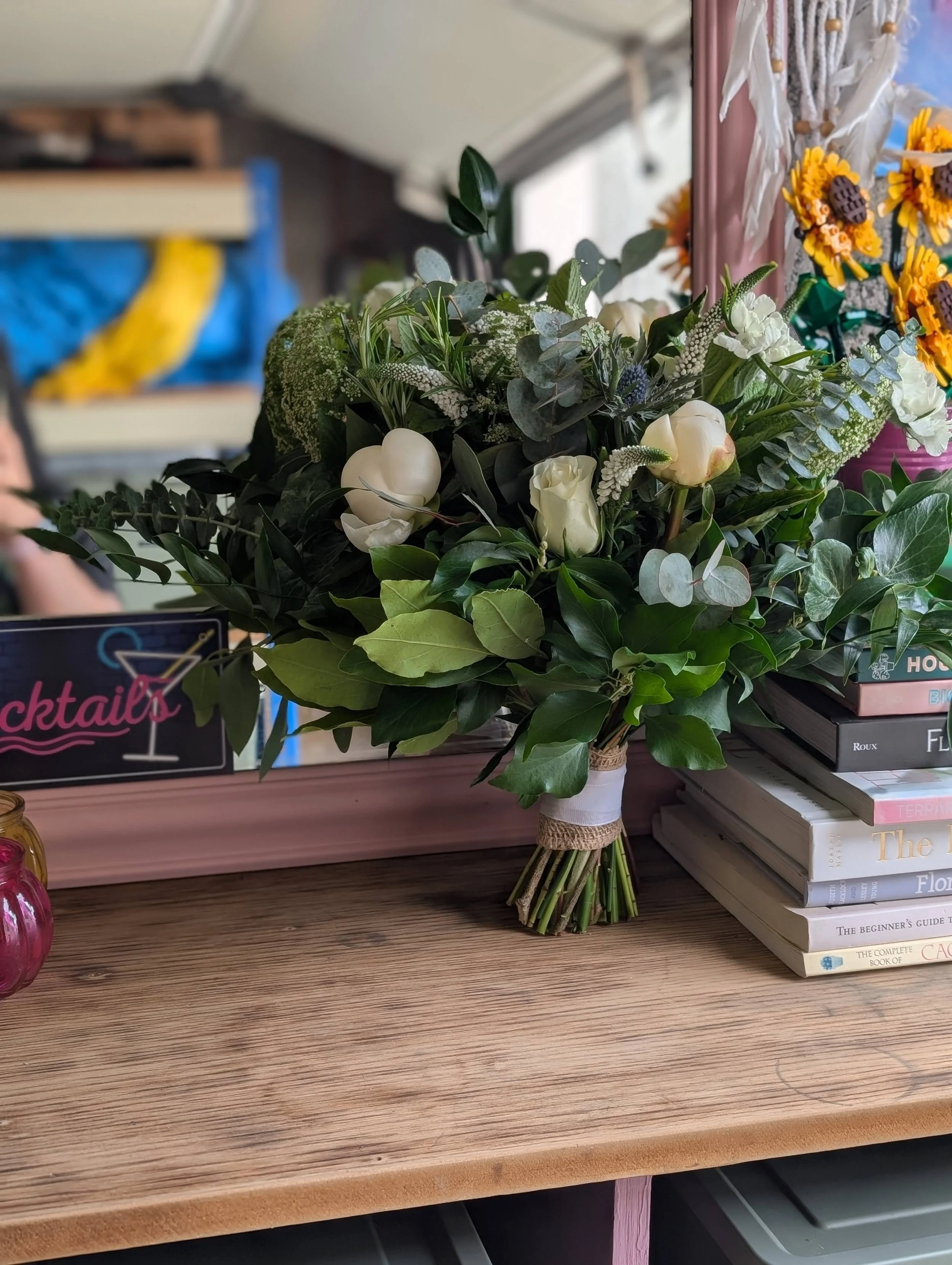 A bouquet of white roses, green leaves, and other greenery tied with a ribbon on a wooden surface, with books and a pink mirror in the background.