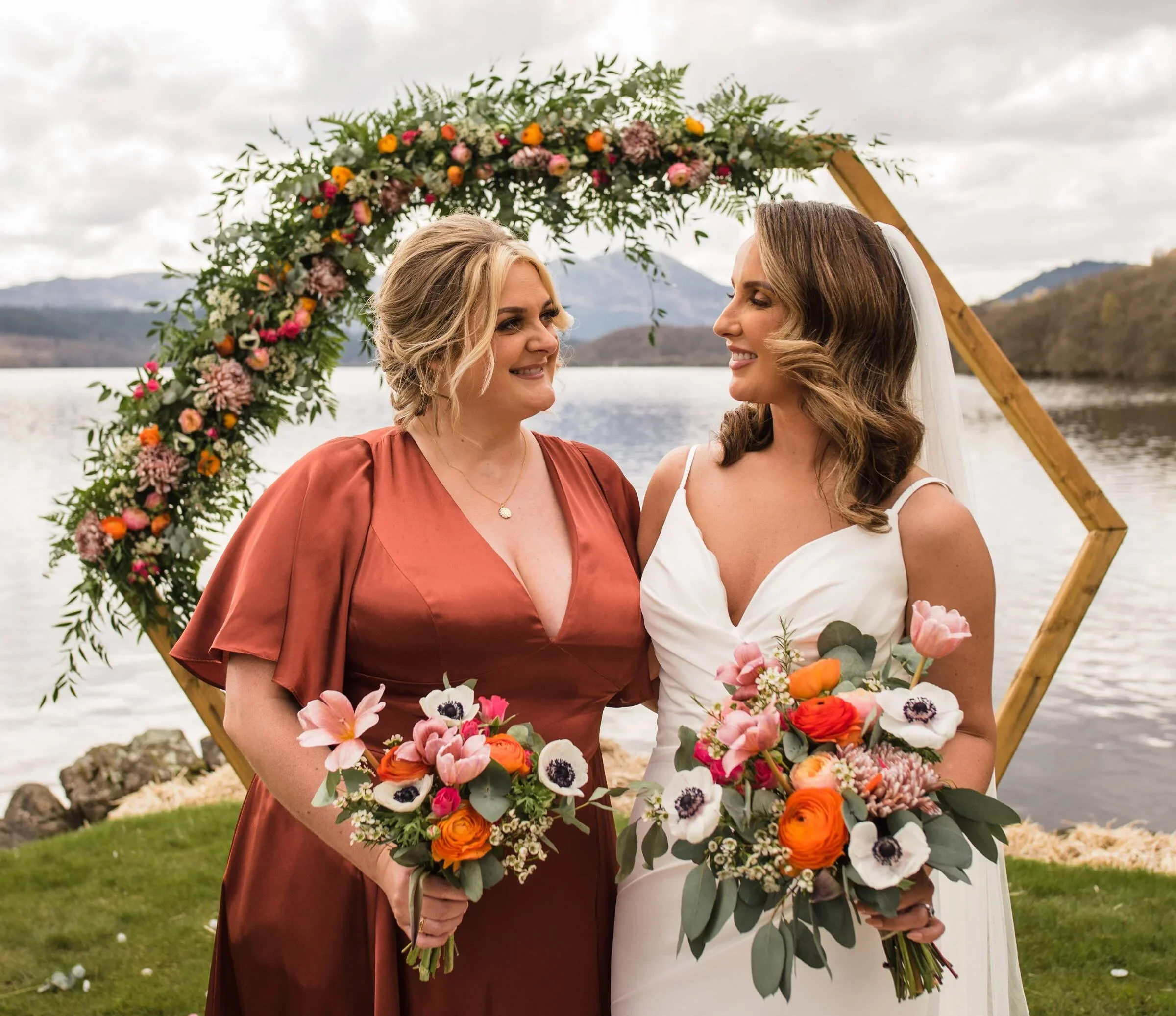 Two women in wedding attire stand facing each other outdoors by a lake with mountains in the background, holding bouquets of colorful flowers.
