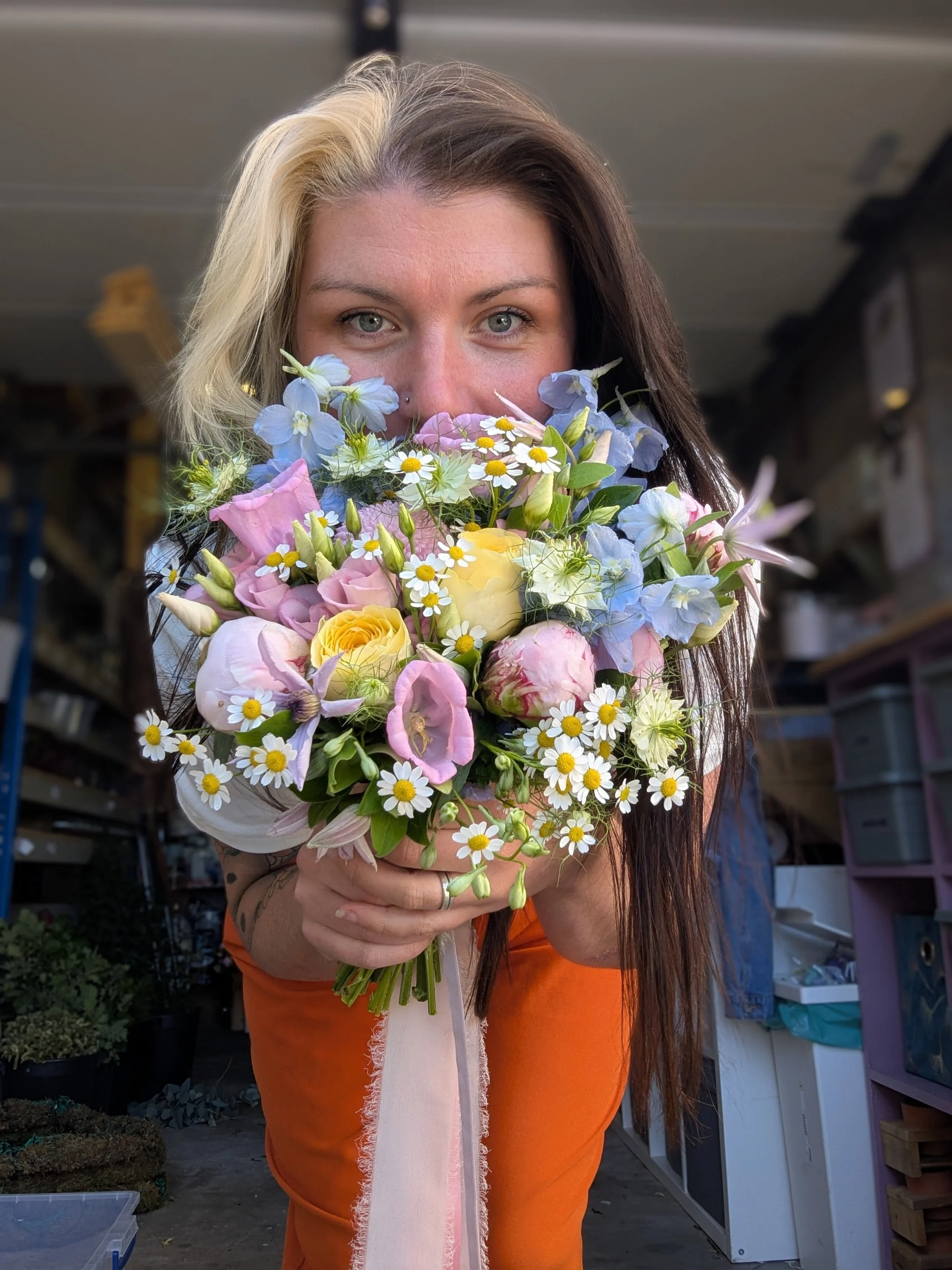 A woman with dark and blonde hair holding a colorful bouquet of flowers close to her face inside a workshop or storage area.
