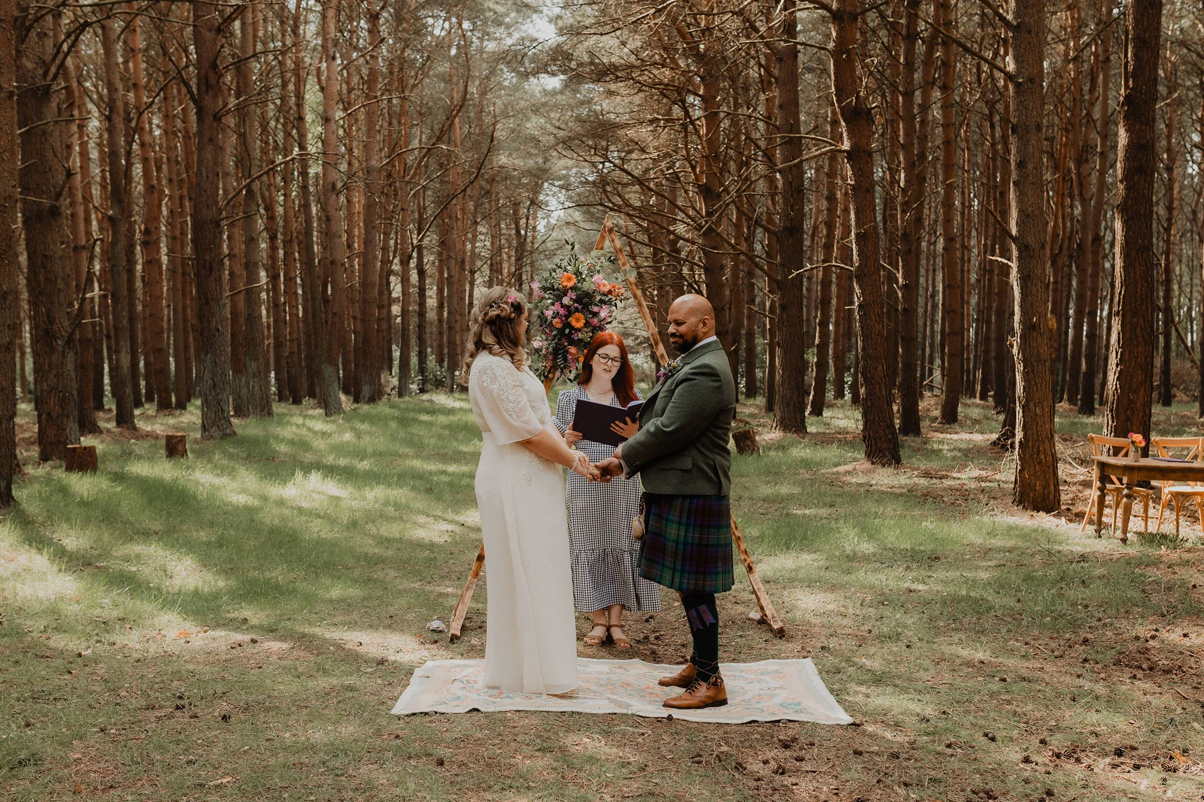 A couple getting married in a forest, holding hands and looking at each other, with an officiant standing in front of them, and a floral arrangement and a wooden triangle decoration behind them.