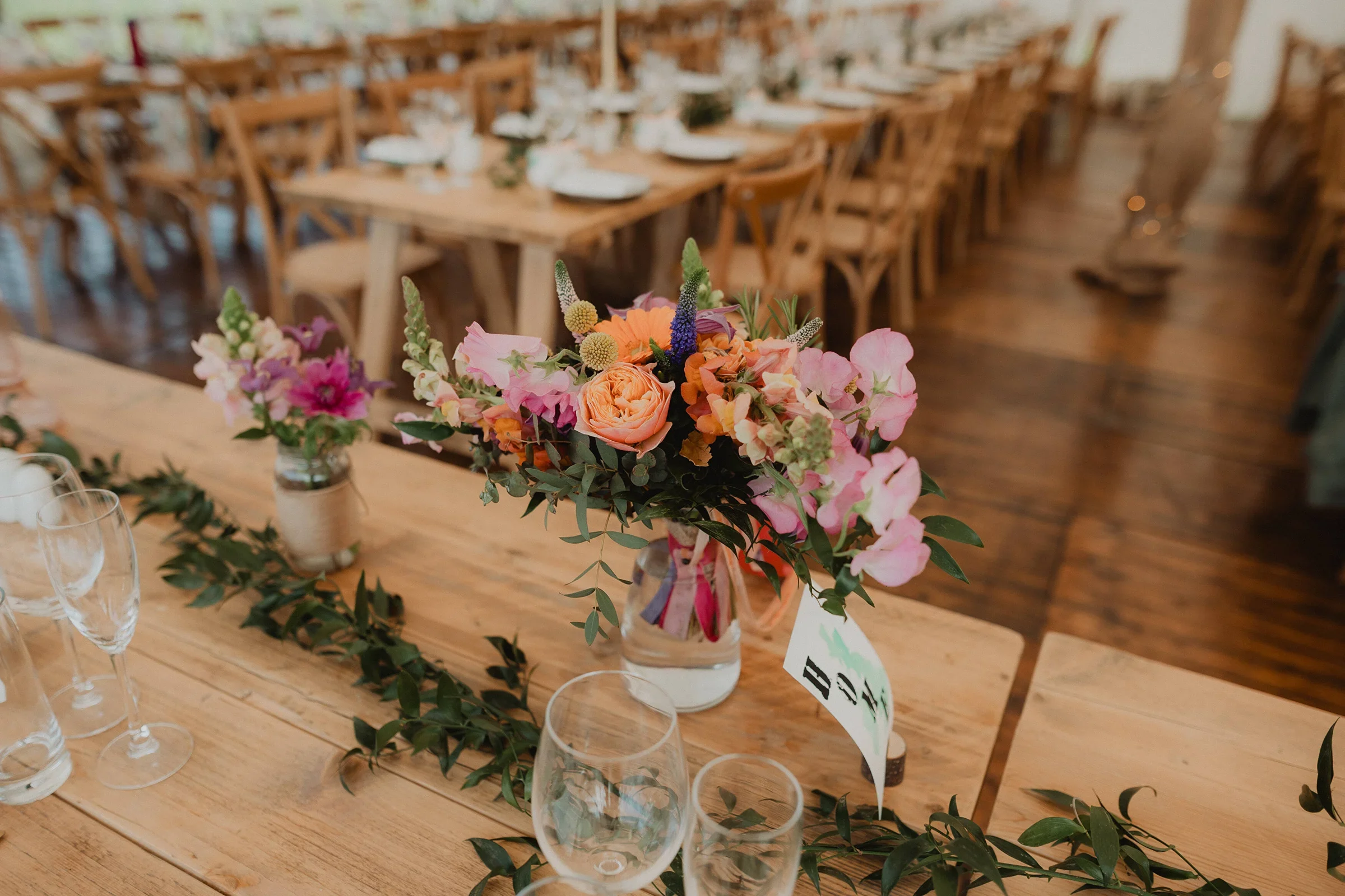 A floral centerpiece featuring pink, peach, and purple flowers on a wooden table at a wedding reception or event, with empty glasses and a table number card nearby.