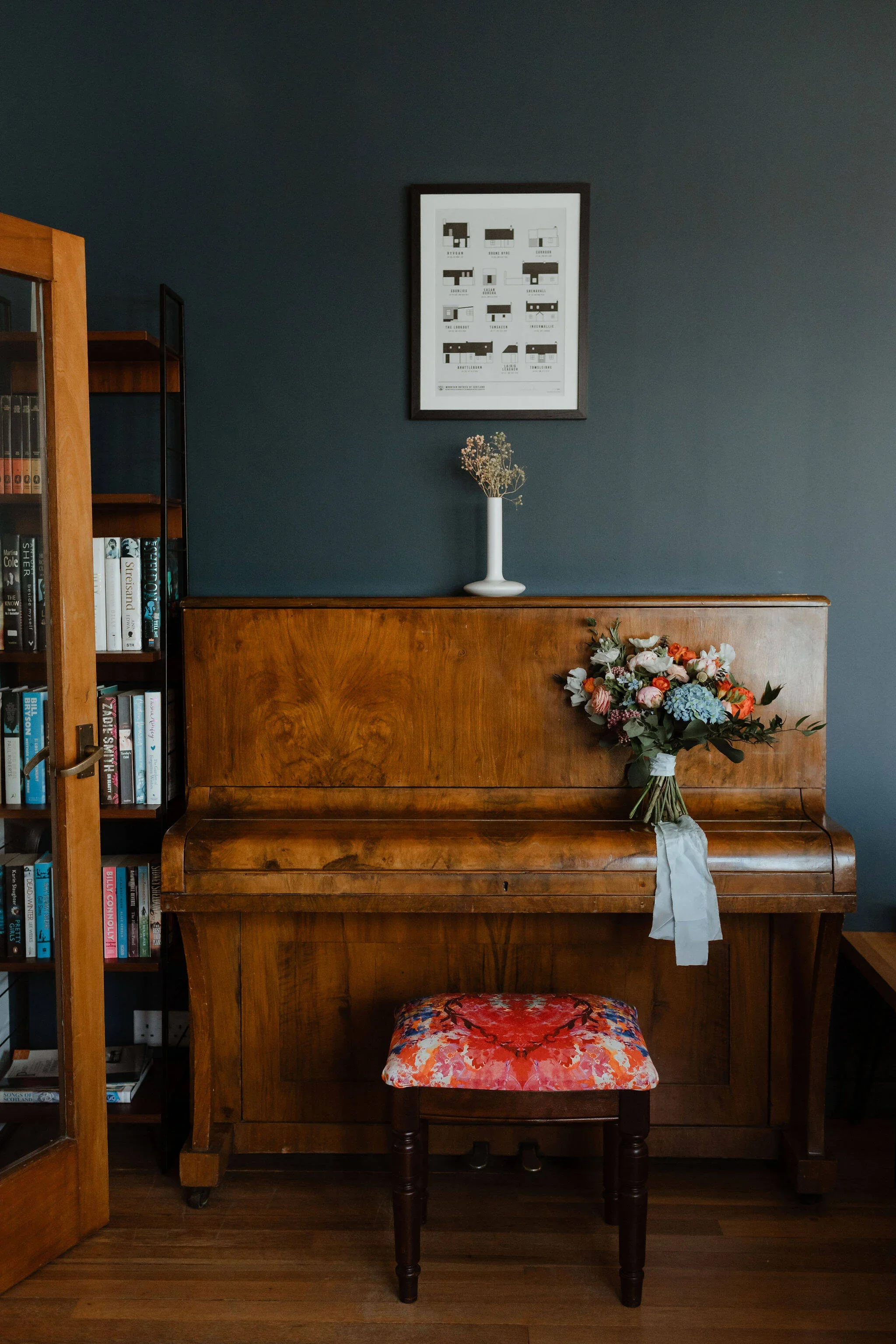 Wooden piano with flowers on top, a framed art piece on a dark blue wall, and a bookshelf to the side.
