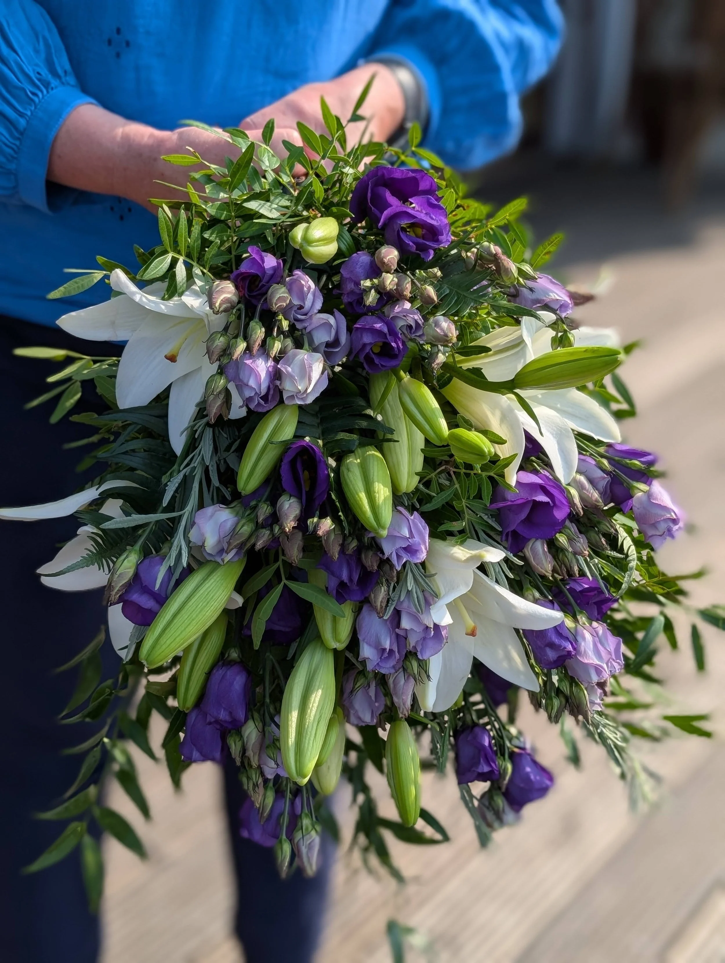 A person in a blue jacket holding a bouquet of white lilies, purple lisianthus, and greenery.
