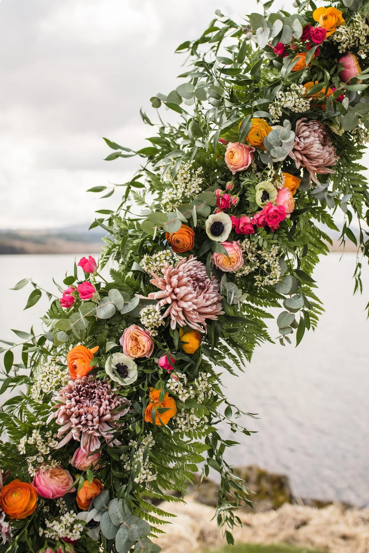 Colorful flower arrangement with pink, orange, white, and yellow flowers and green foliage, hanging outdoors near a body of water.