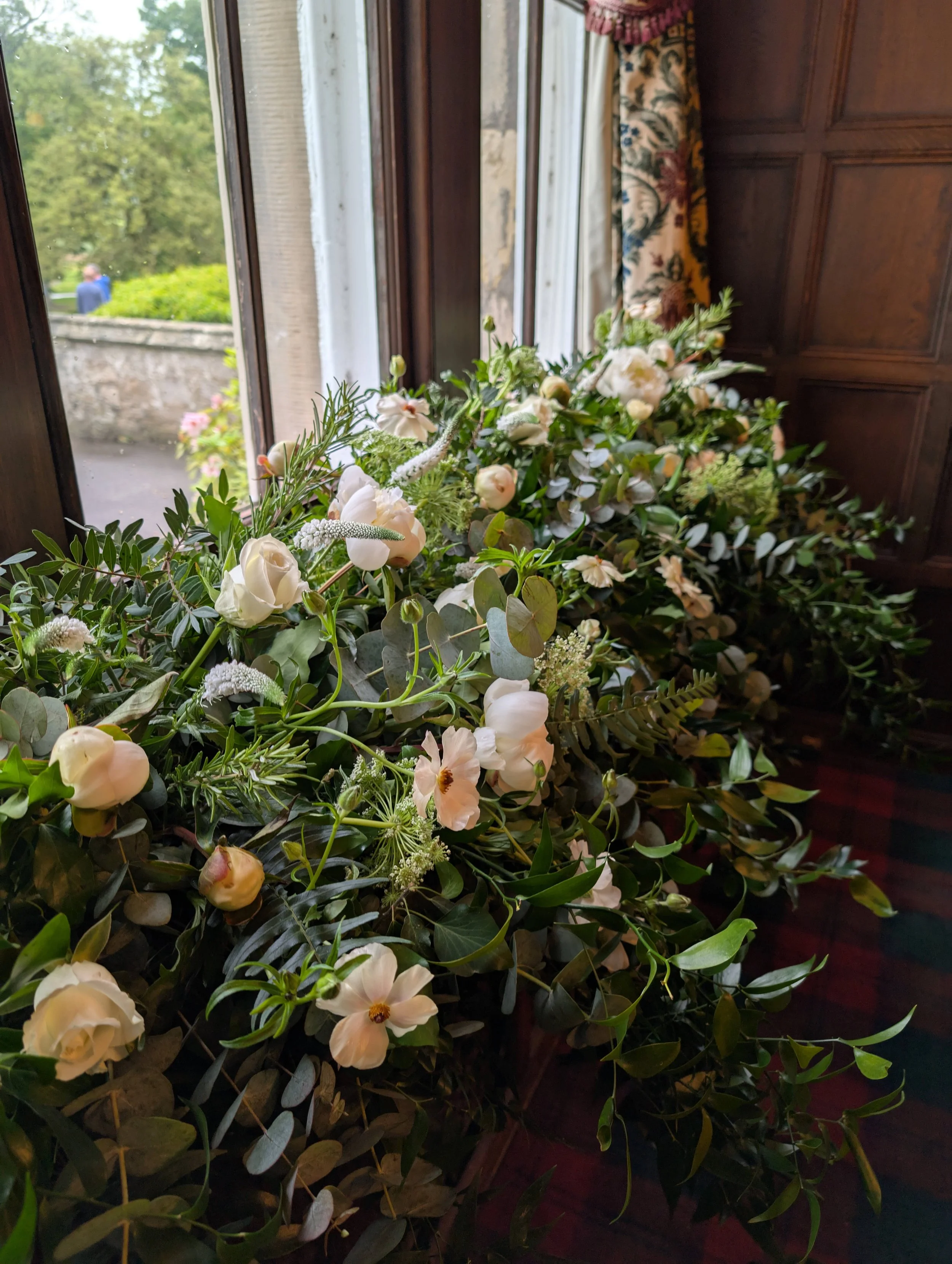 A large floral arrangement of white flowers and green foliage resting in front of a window with floral curtains in a wooden-paneled room.