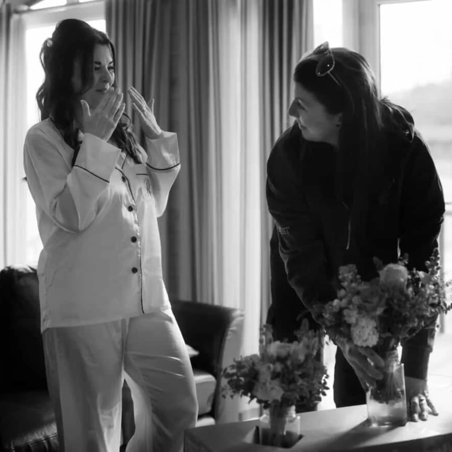 Two women, one in pajamas, reacting happily as the other presents a bouquet of flowers in a living room with windows and curtains.