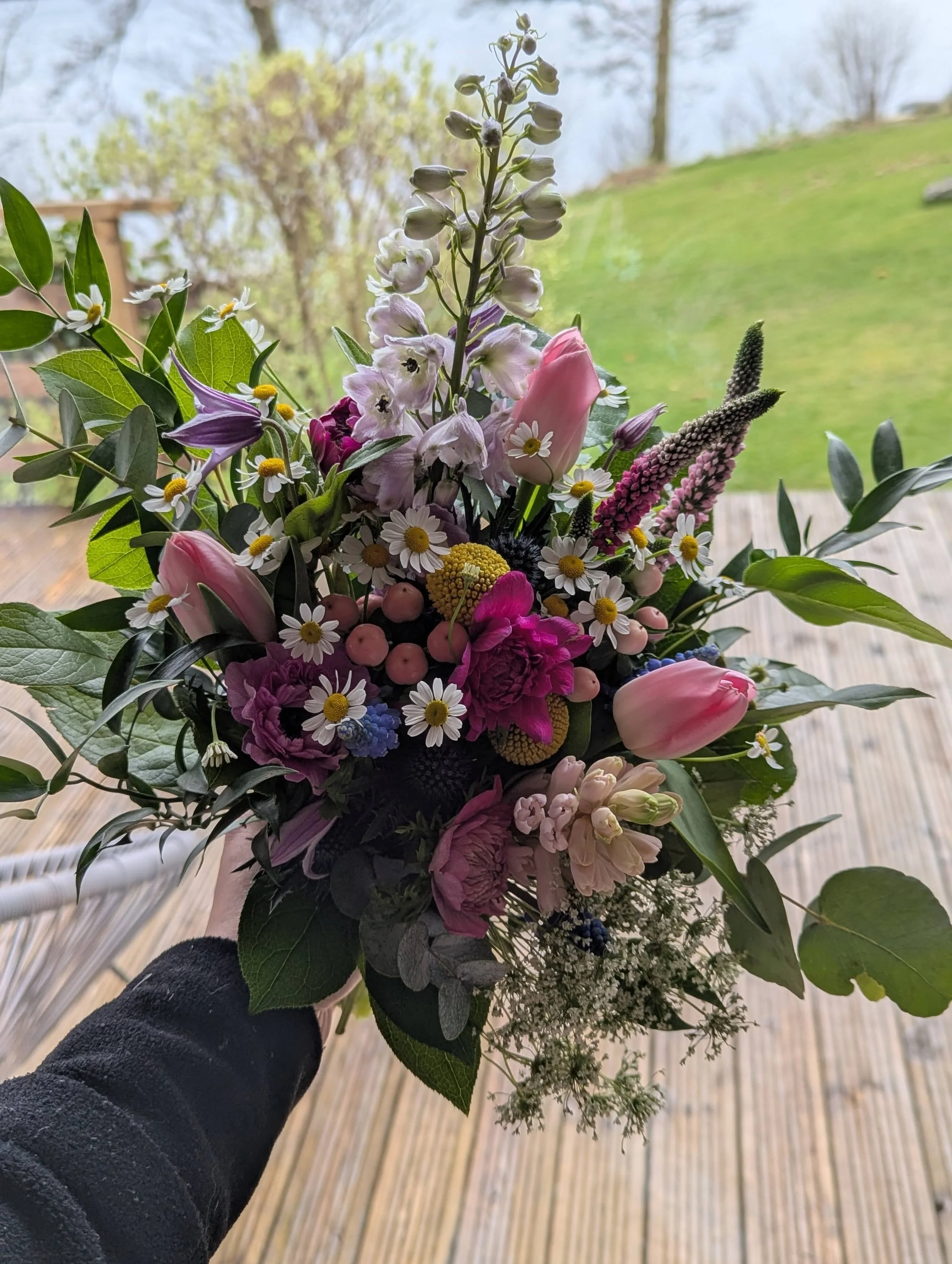 A colorful bouquet of mixed flowers including pink tulips, white daisies, purple carnations, and other small flowers held outdoors on a wooden deck with a green lawn and trees in the background.