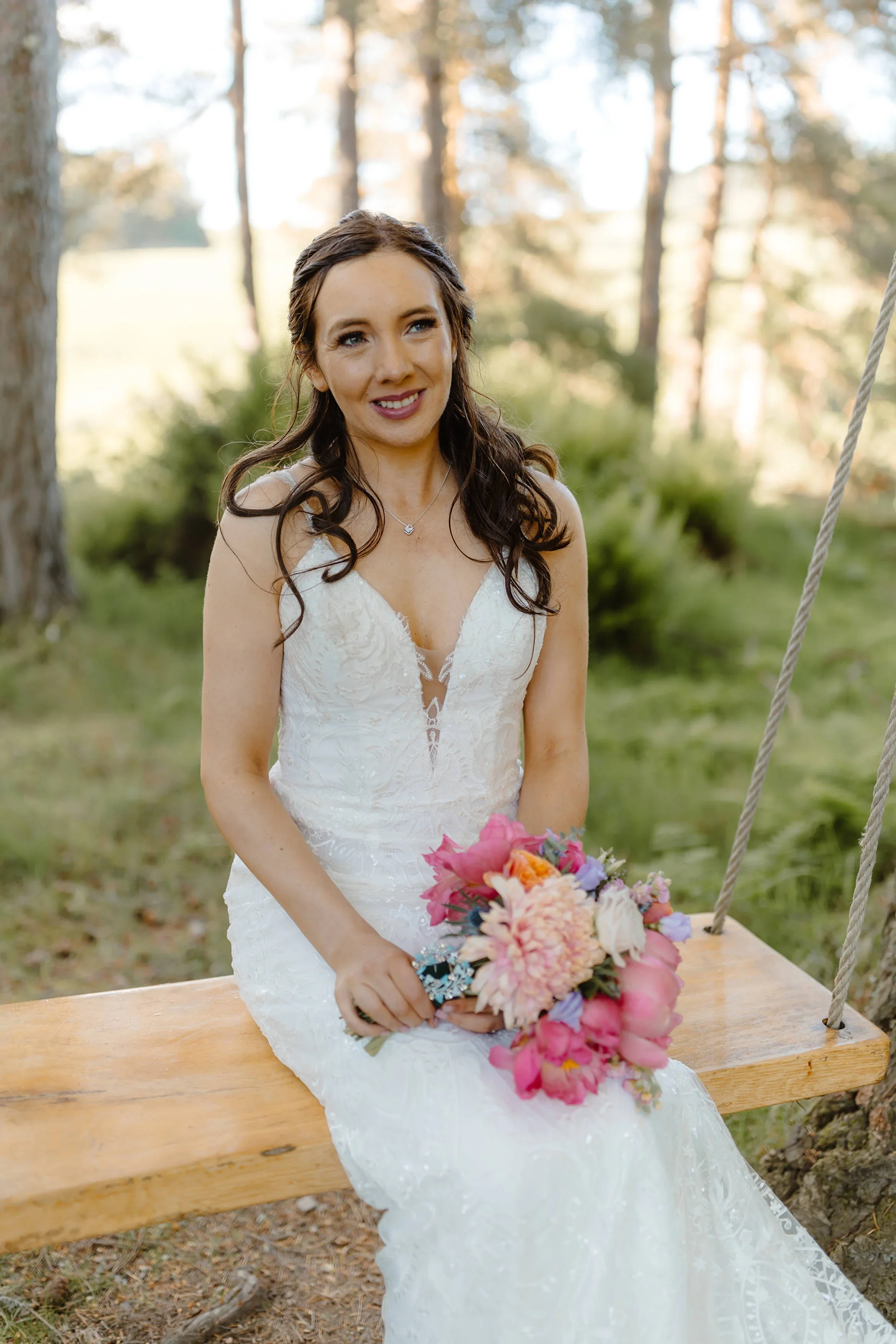A woman in a white lace wedding dress sitting on a wooden swing outdoors, holding a colorful bouquet of flowers, with trees and green foliage in the background.