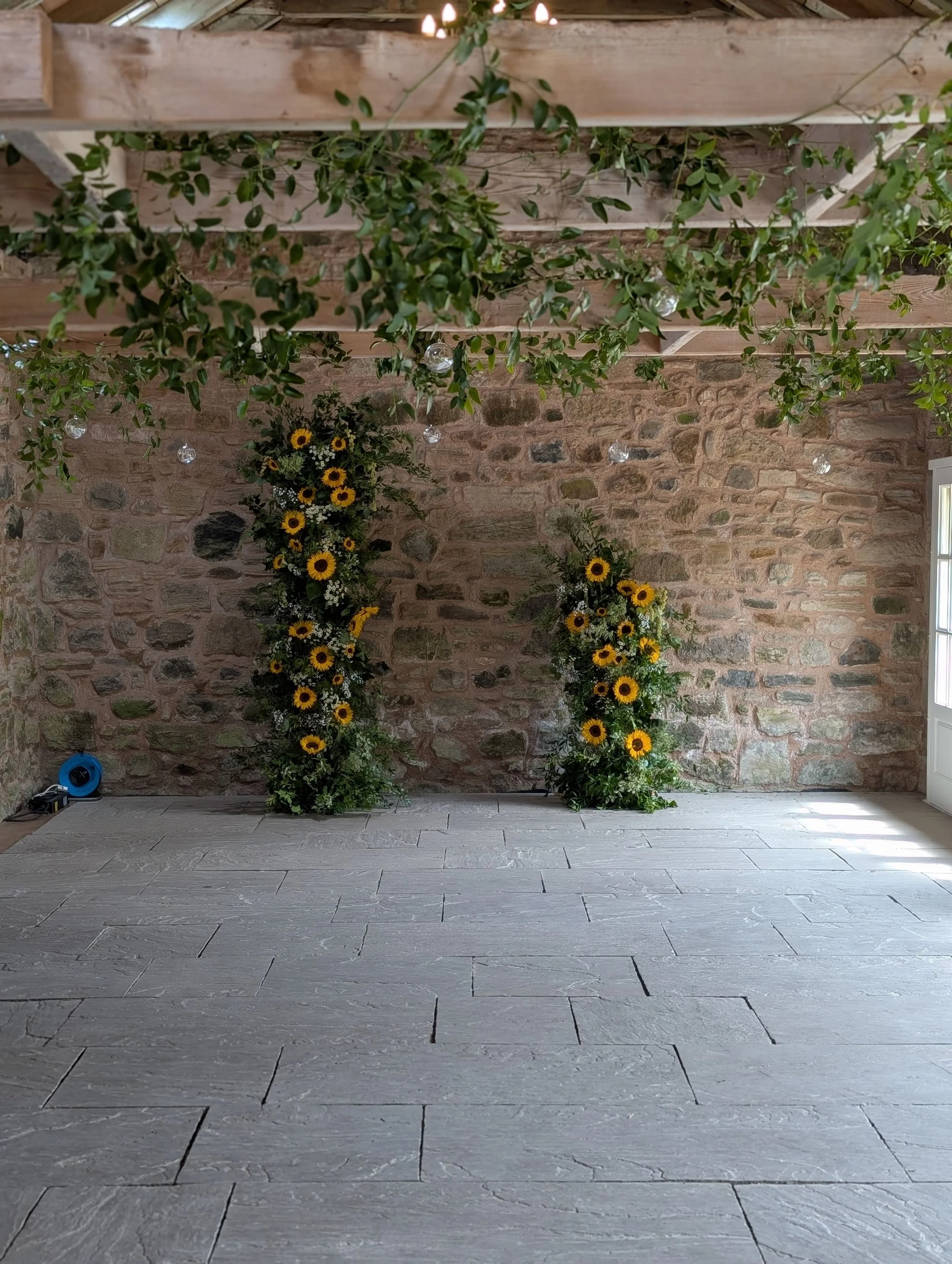 Indoor space with a brick wall background, decorated with two floral arrangements of sunflowers and greenery, and a wooden ceiling with hanging greenery and small hanging lights.