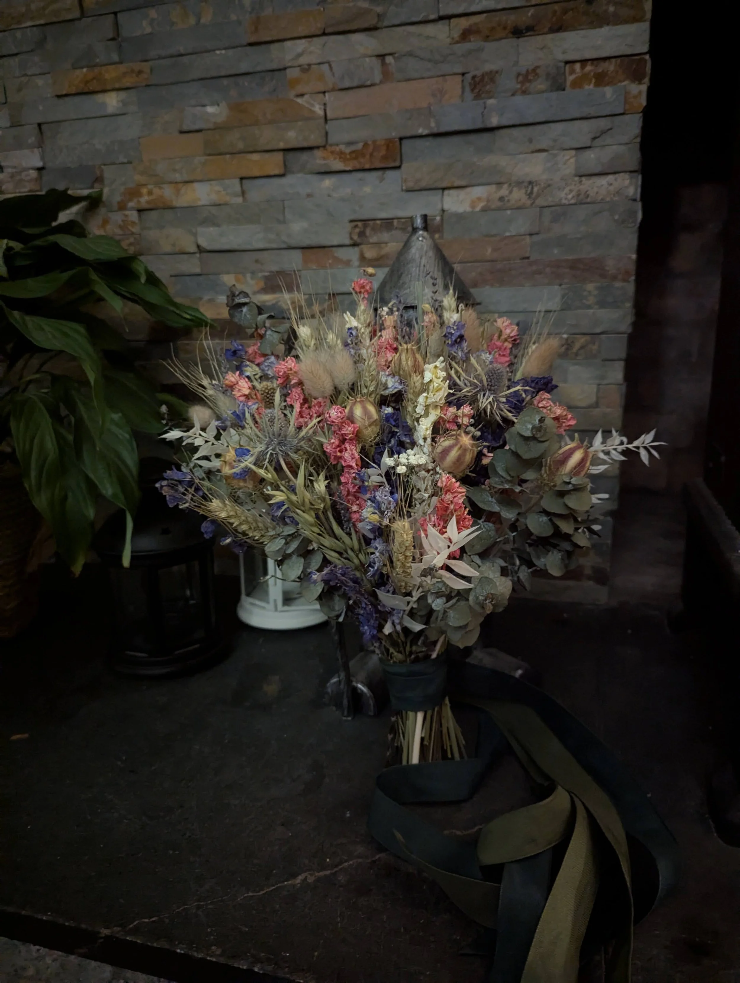 A bouquet of dried flowers including purple, pink, and white flowers, with greenery and seed pods, placed on a dark surface against a stone wall background.