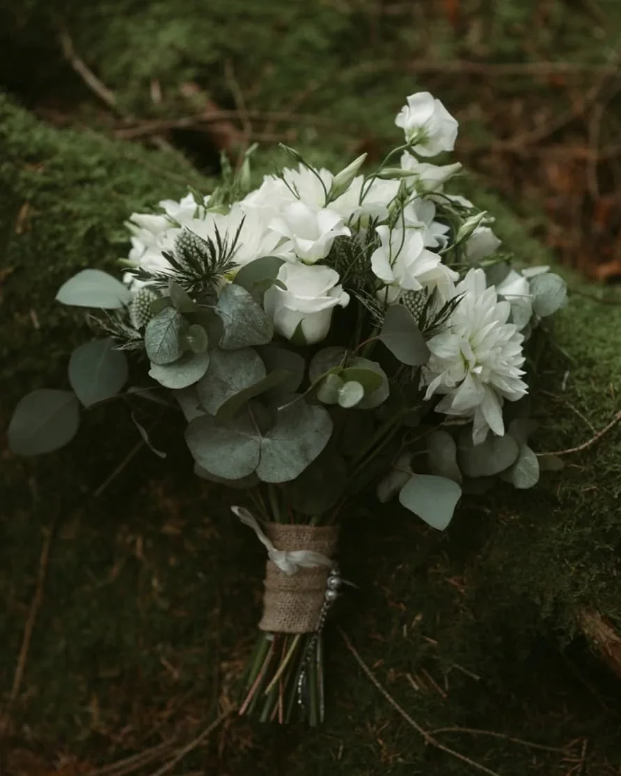 A bouquet of white flowers and greenery wrapped with burlap and tied with string, resting on forest floor moss and twigs.