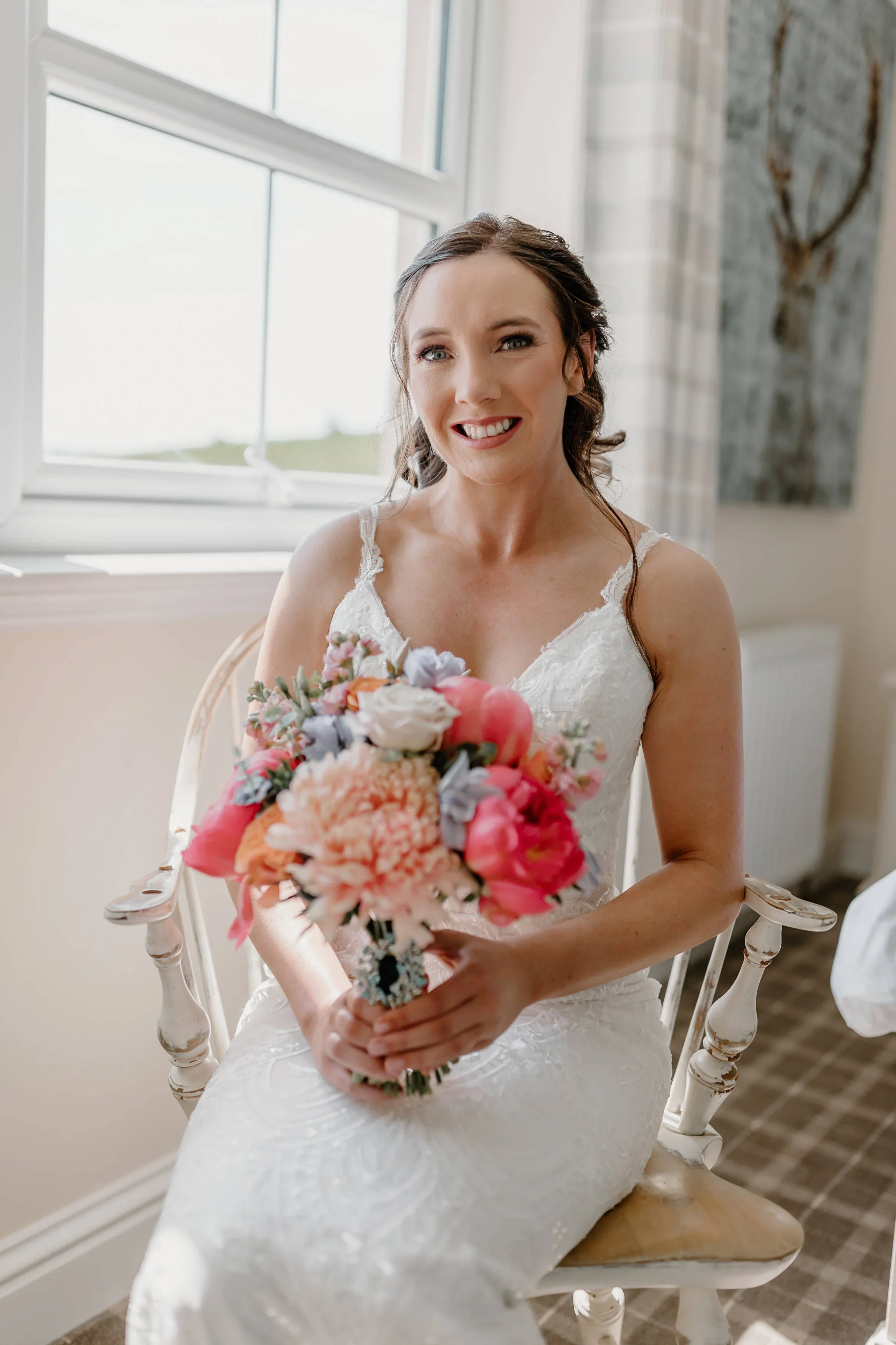 A smiling bride in a white wedding dress sitting on a chair by a window, holding a bouquet of pink, white, and purple flowers.