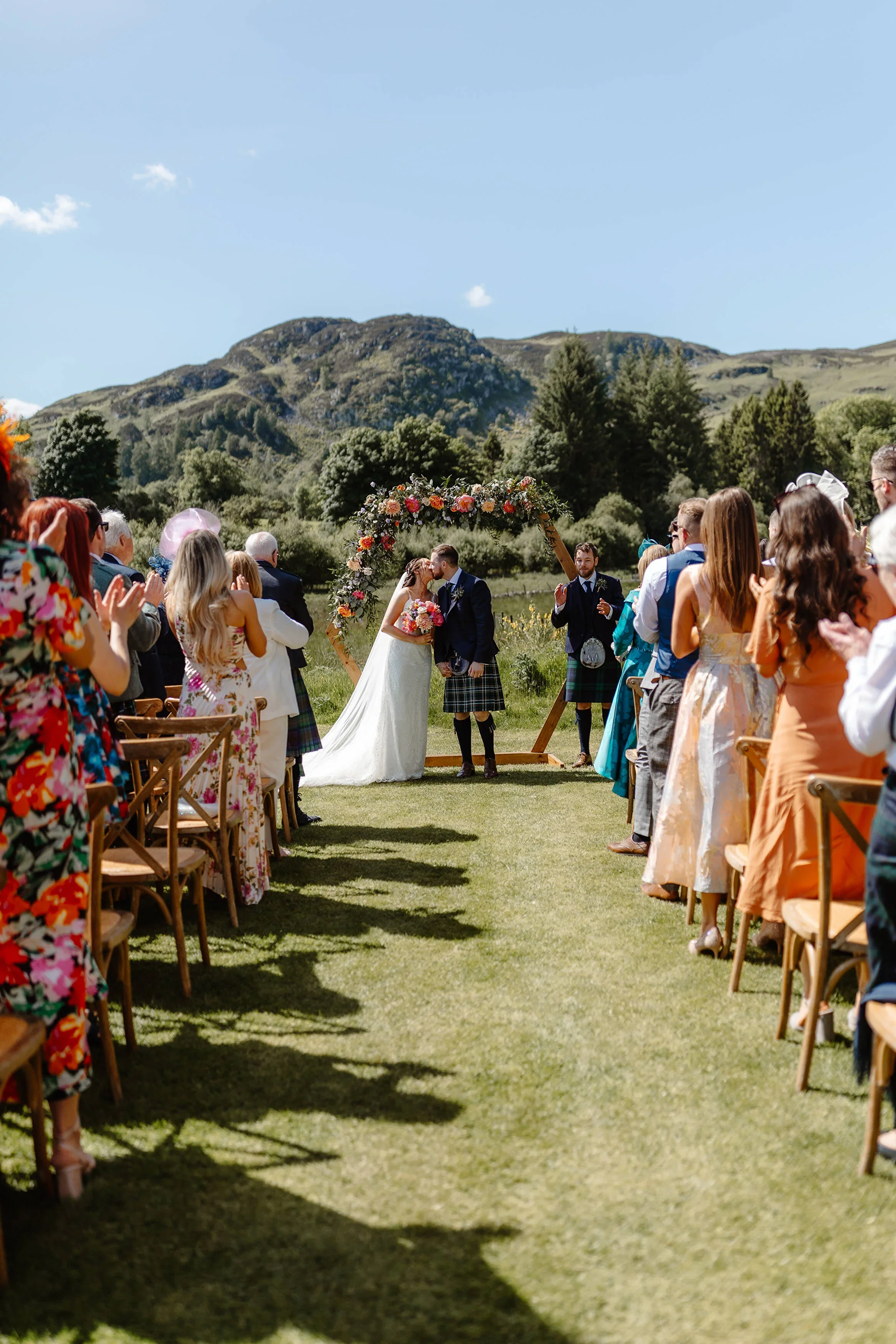 A wedding ceremony outdoors with the bride and groom sharing a kiss under a floral arch, surrounded by guests clapping on a grassy field with mountains in the background.