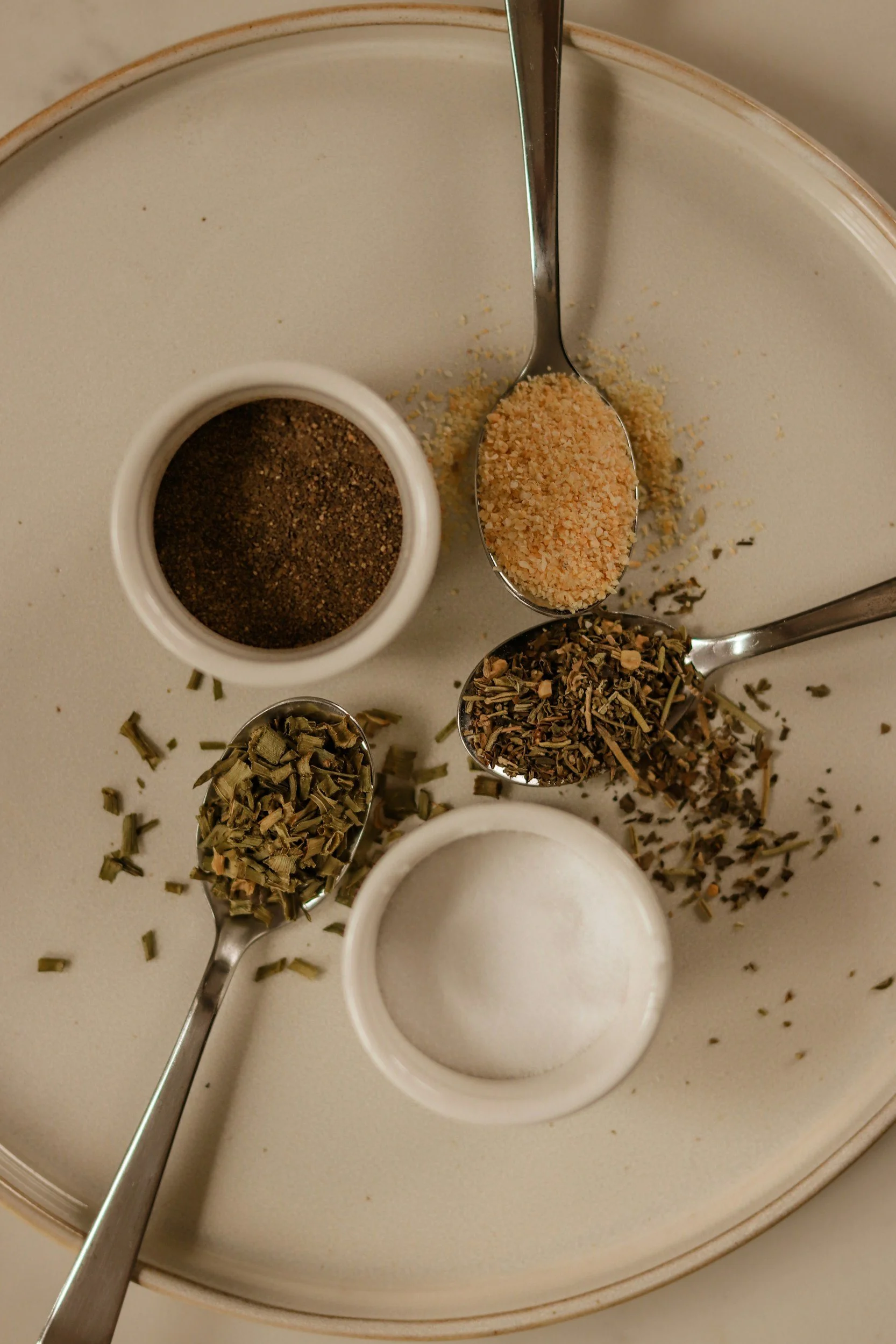 Dried spices and herbs in spoons and small containers on a plate, including ground black pepper, salt, garlic powder, and aromatic herbs.