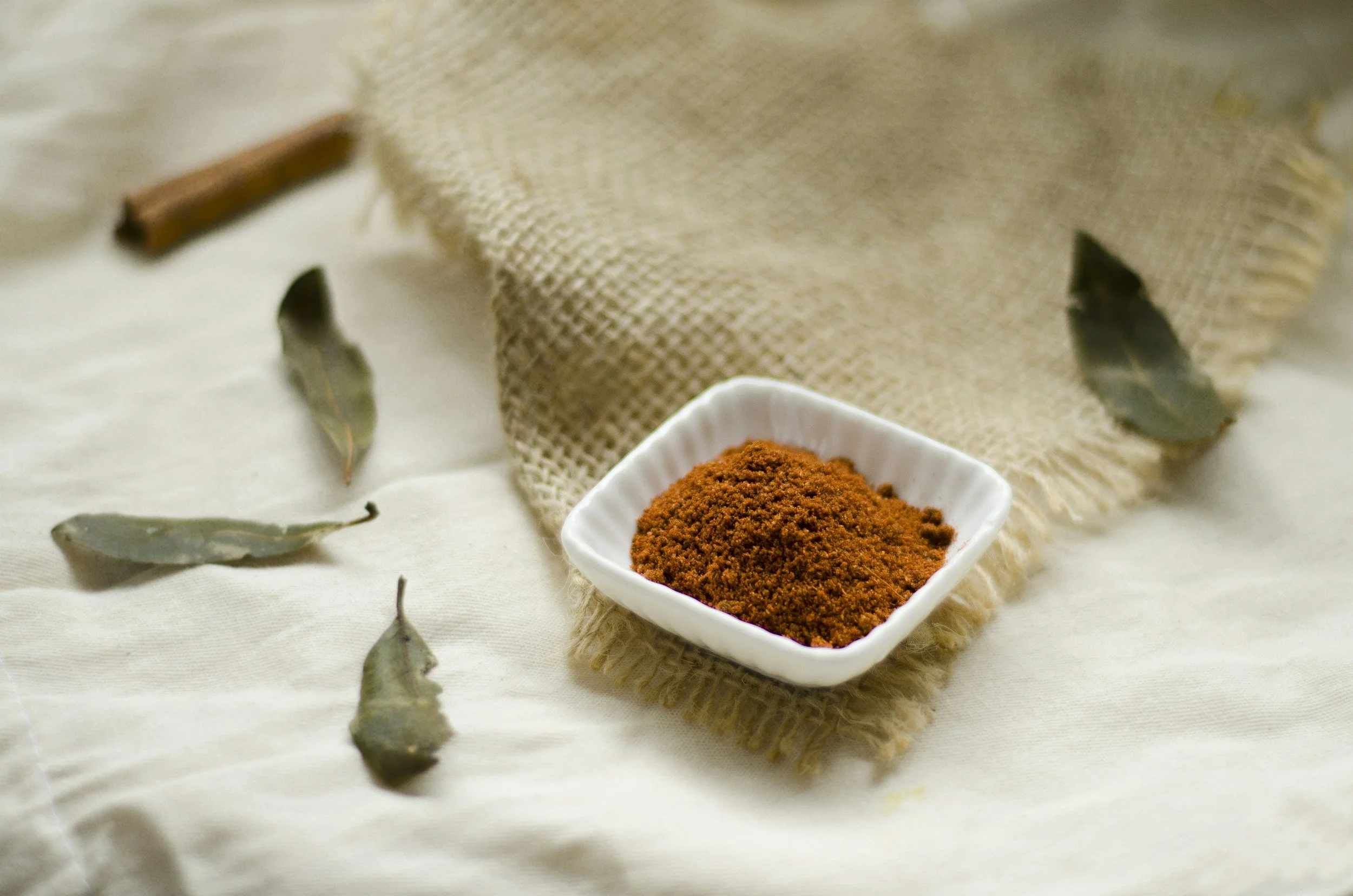 Red spice powder on a white plate with dried bay leaves, cinnamon sticks, and burlap fabric in the background.