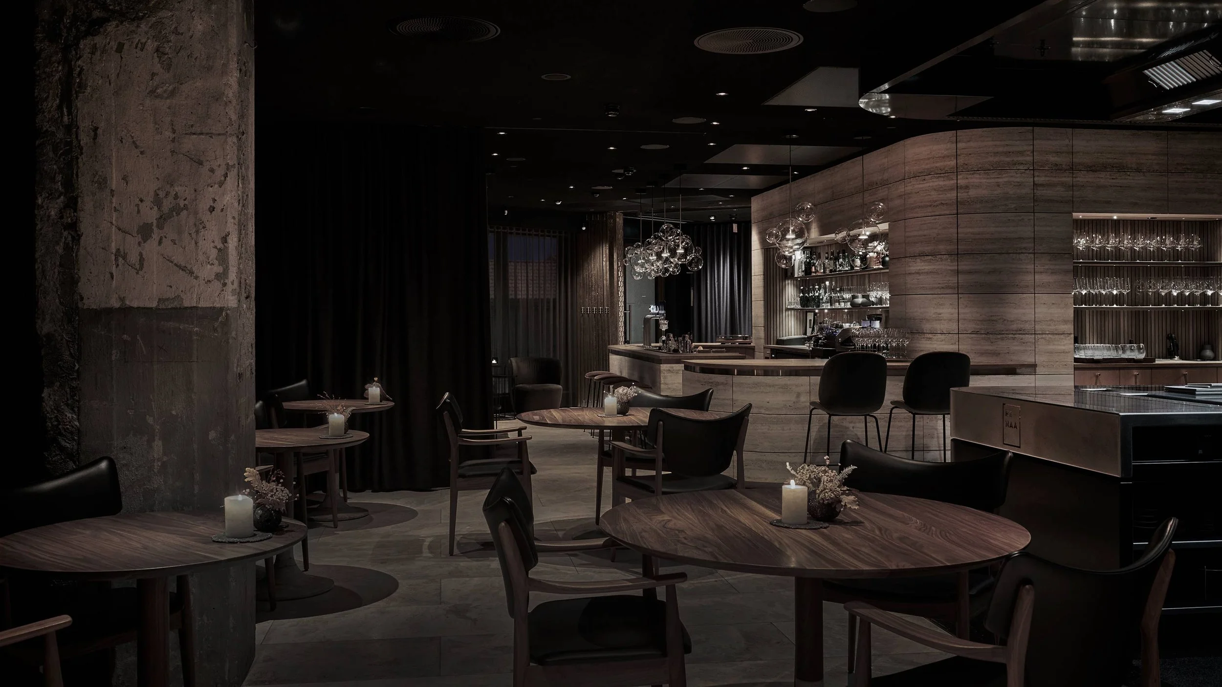 A dimly lit modern restaurant or bar with wooden tables, chairs, a curved bar counter, glass shelves displaying glassware, decorative hanging glass orbs, and small lit candles on tables.