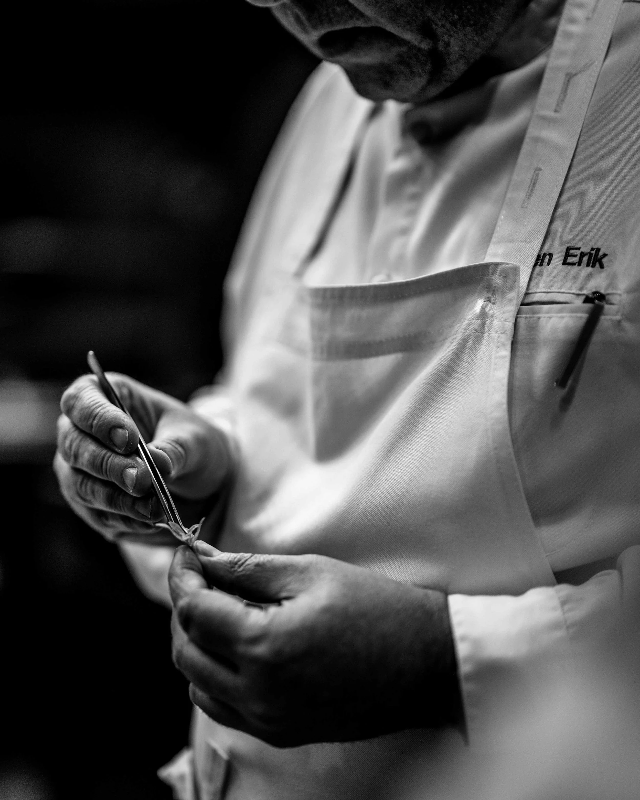 Black and white photo of a person in a white coat holding a small surgical instrument and a thread, possibly performing a medical or surgical procedure.