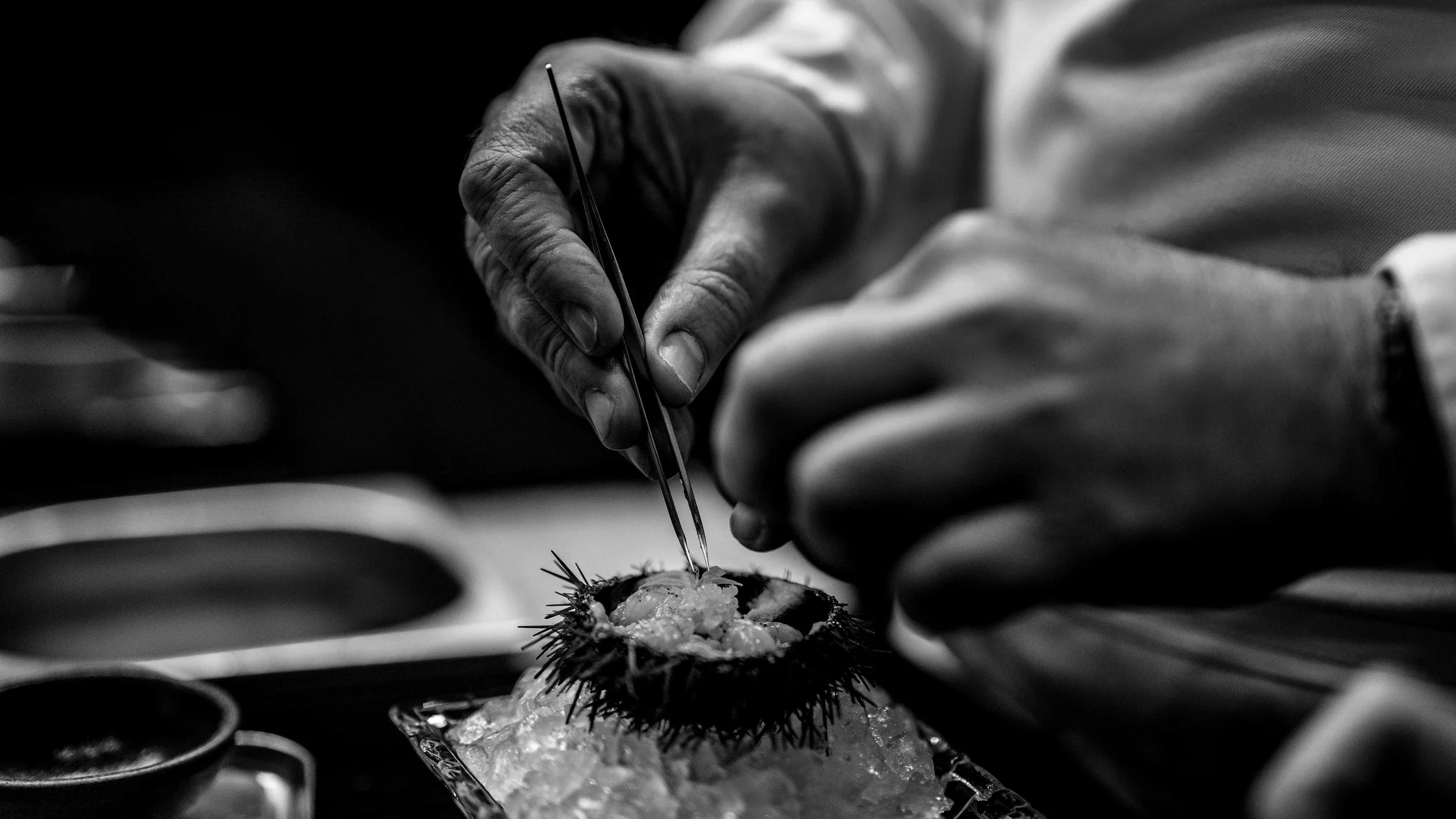 A person using tweezers to prepare a sea urchin for eating, with sushi dishes in the background in black and white.