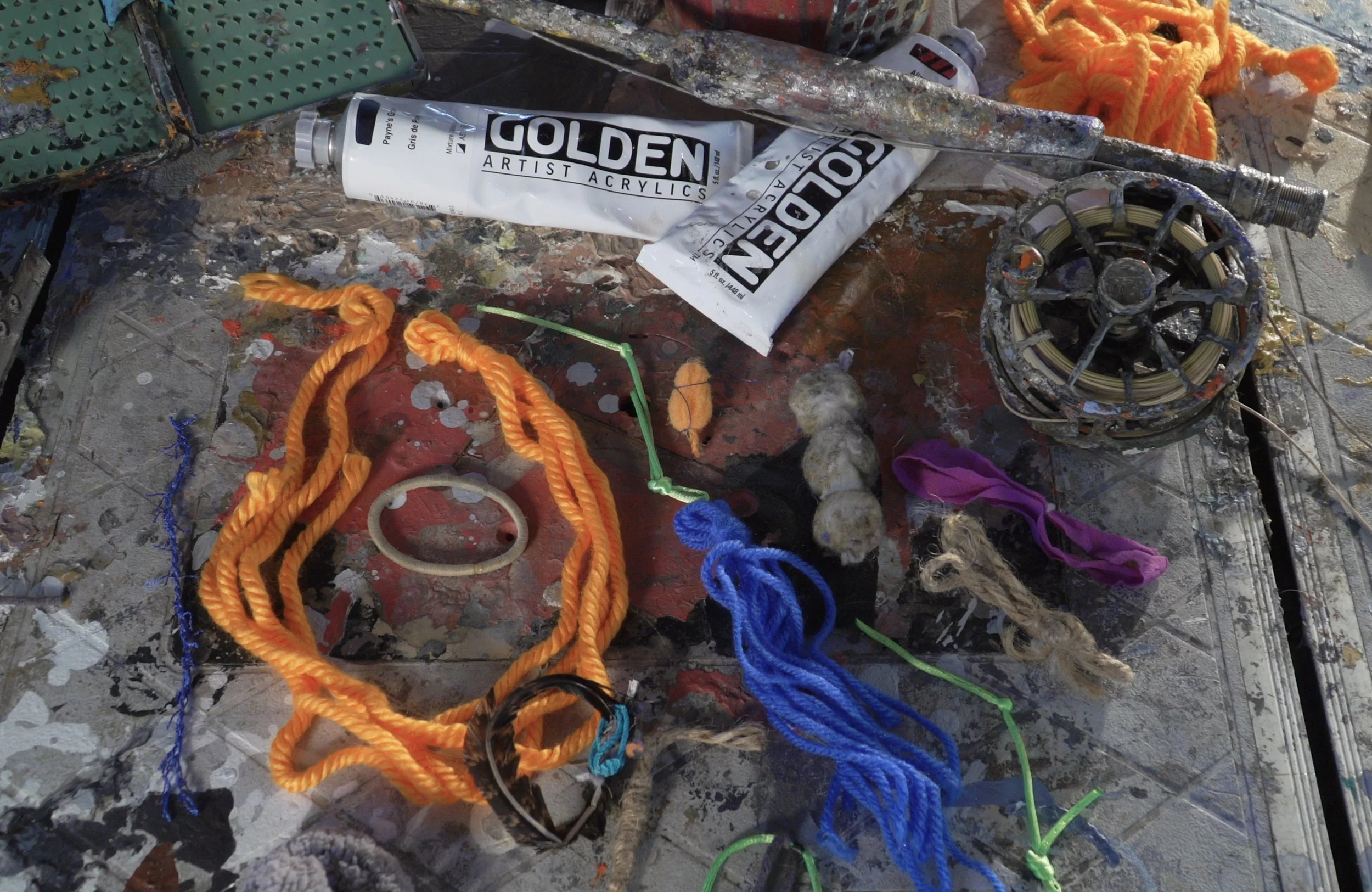 An artist's work surface with various art supplies, including tubes of Golden acrylic paint, colorful ropes, a small white ring, a spool, and several pieces of fabric or yarn in different colors. The surface is splattered with paint.