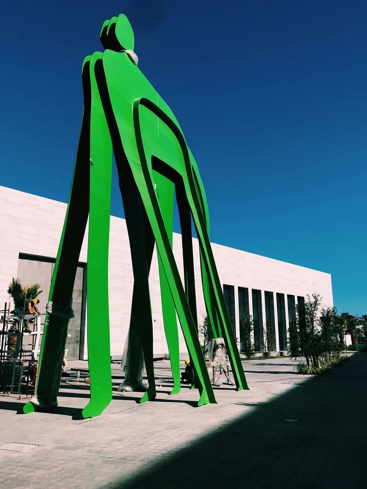 Large green abstract sculpture with black accents outside a modern white building with tall windows, under a clear blue sky.