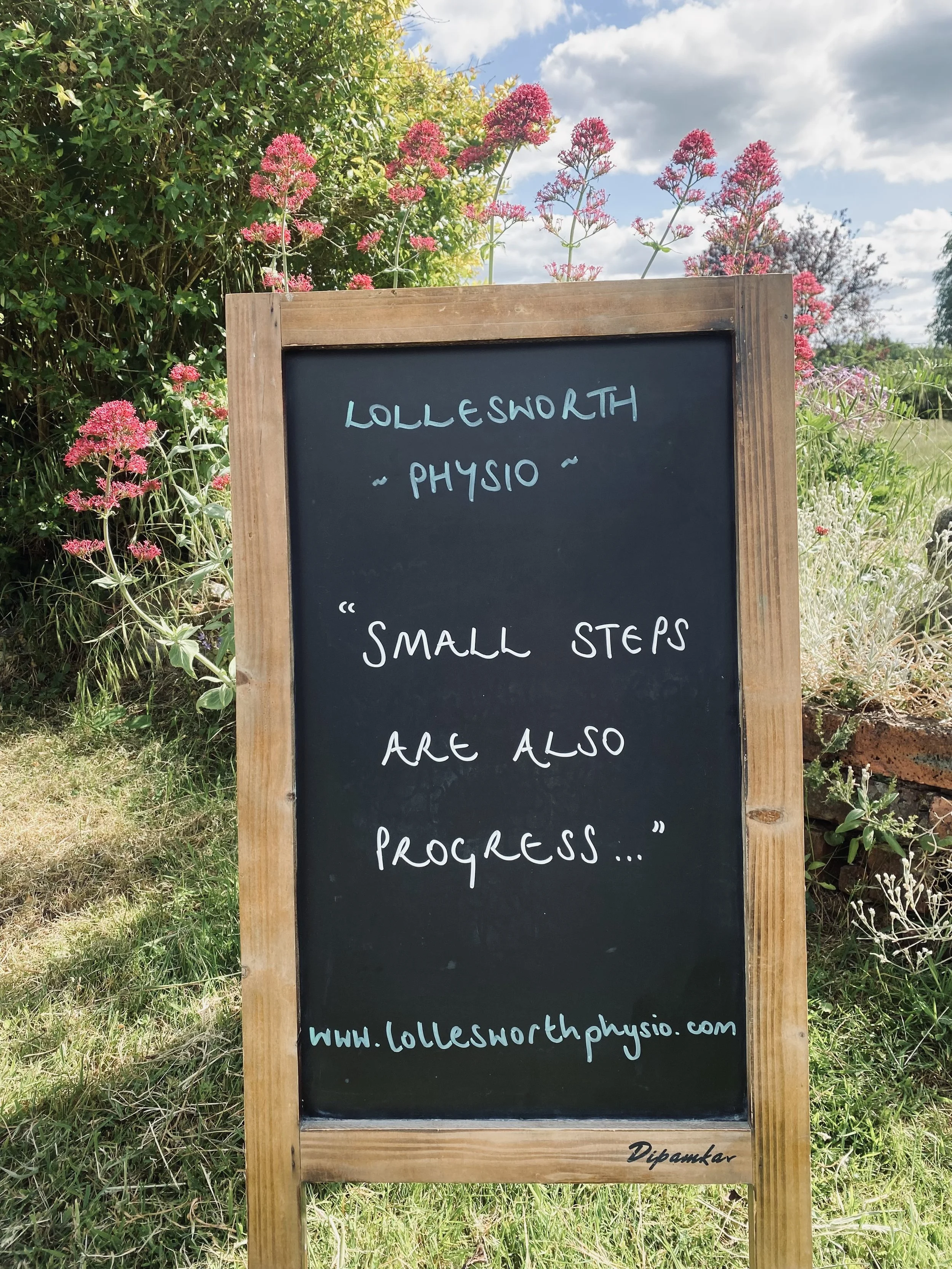 A black chalkboard sign with a wooden frame outdoors, surrounded by pink and purple flowers and green foliage, with a partly cloudy sky in the background. Horsely, Surrey.