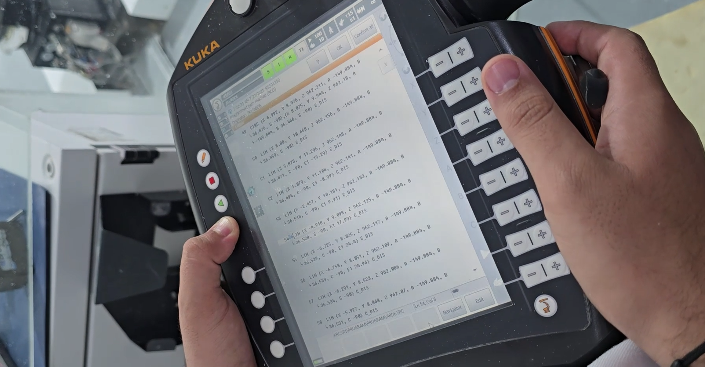 Cep telefonuna bağlı taşıma cihazı, ekranda çeşitli teknik veriler ve kodlar gösteriliyor, kullanıcının sağ el ile kullanımda, arka planda mekanik veya teknik cihazlar var.