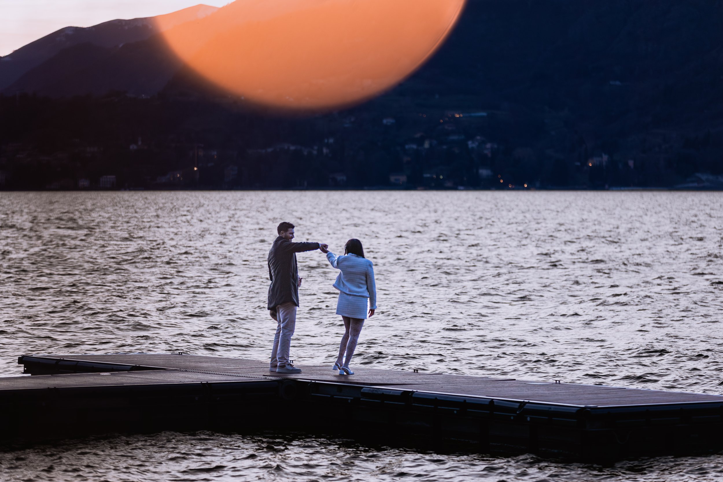 due persone che si tengono per mano sul molo al tramonto, con una montagna sullo sfondo e un grande oggetto rotondo nel cielo