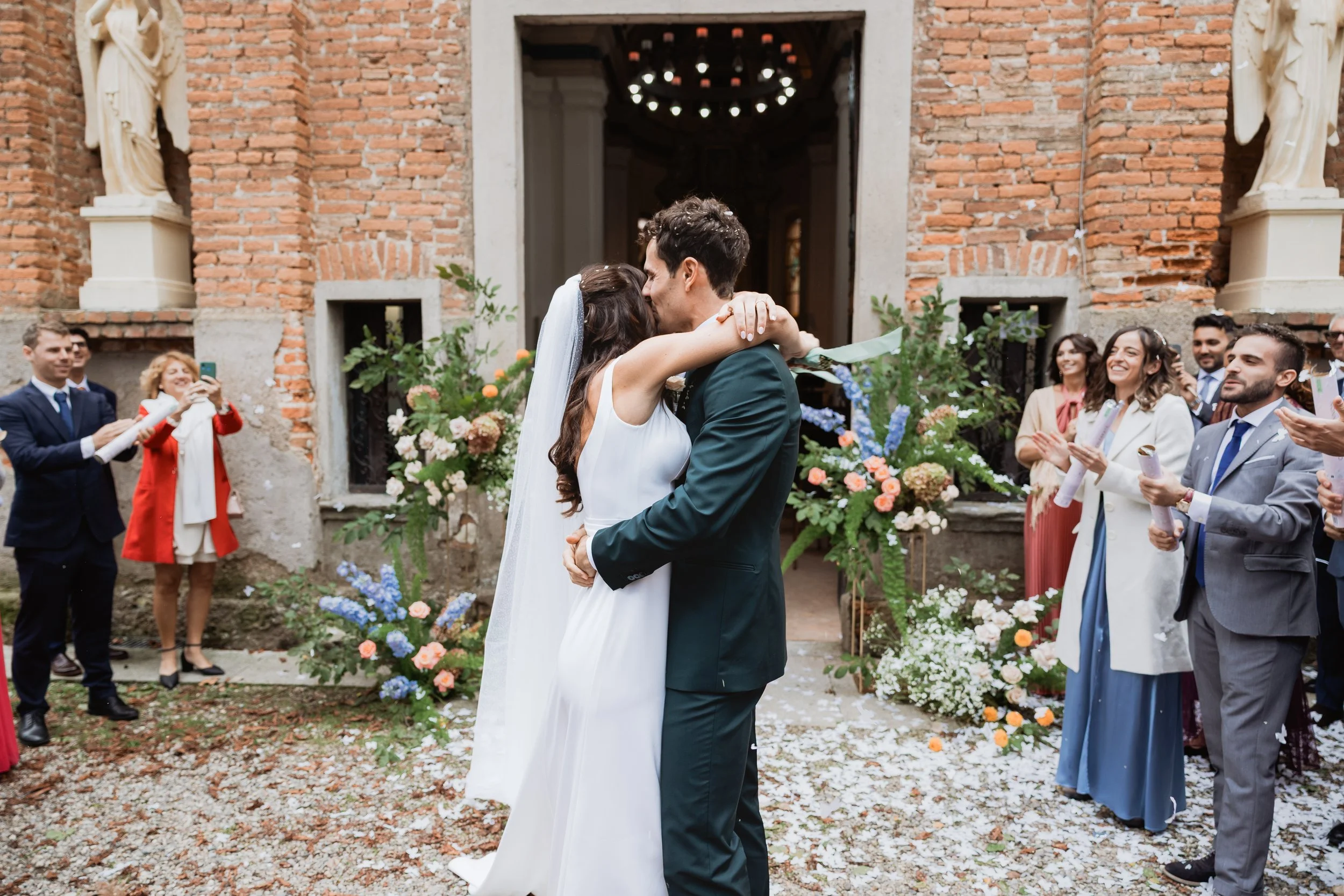 Una coppia di sposi si bacia mentre circondati da amici e parenti in un cortile di chiesa decorato con fiori colorati.