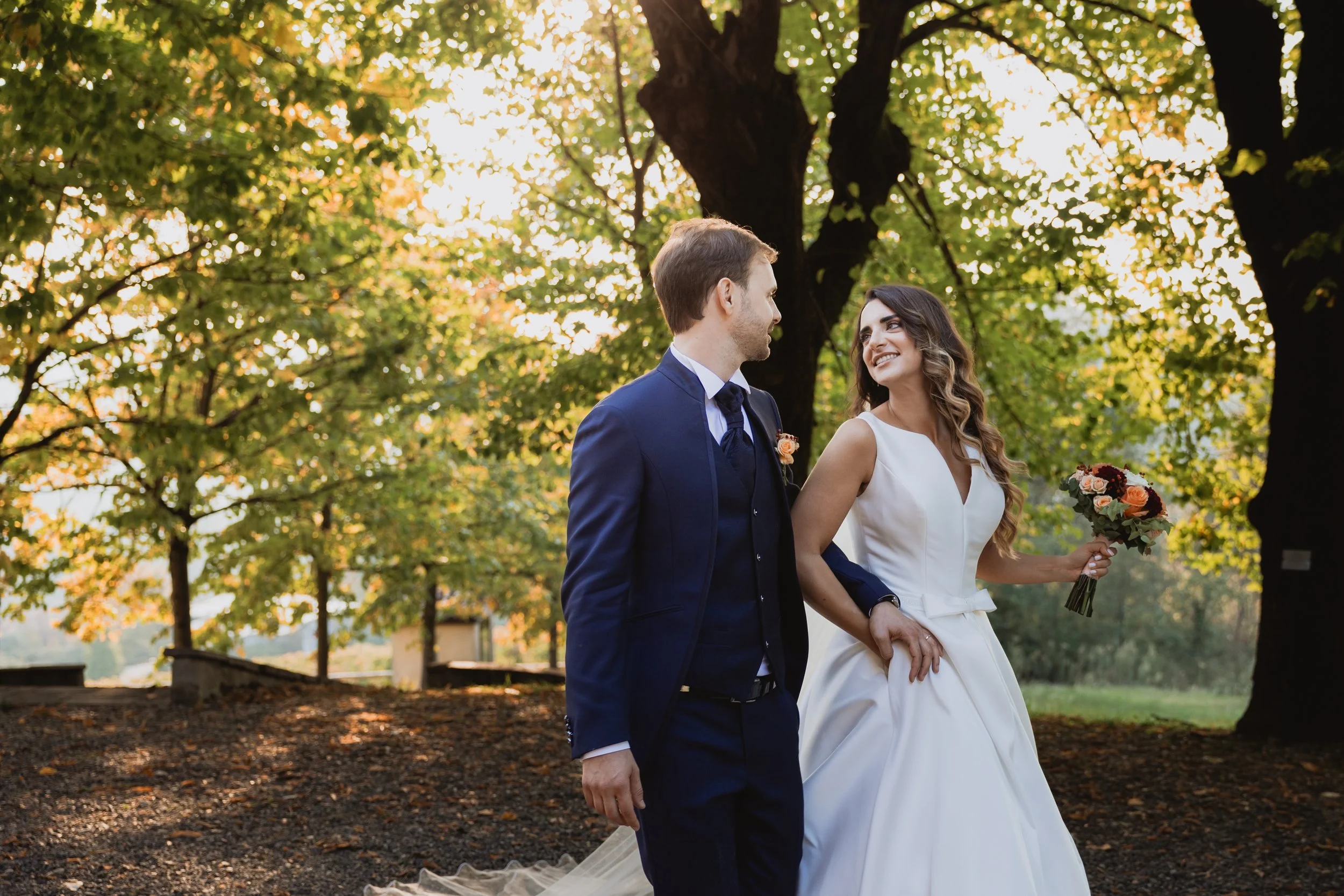 Una coppia di sposi cammina in un parco durante il tramonto, con alberi verdi e foglie autunnali sullo sfondo. La donna indossa un abito da sposa bianco e tiene un bouquet di fiori, mentre l'uomo indossa un completo blu scuro.