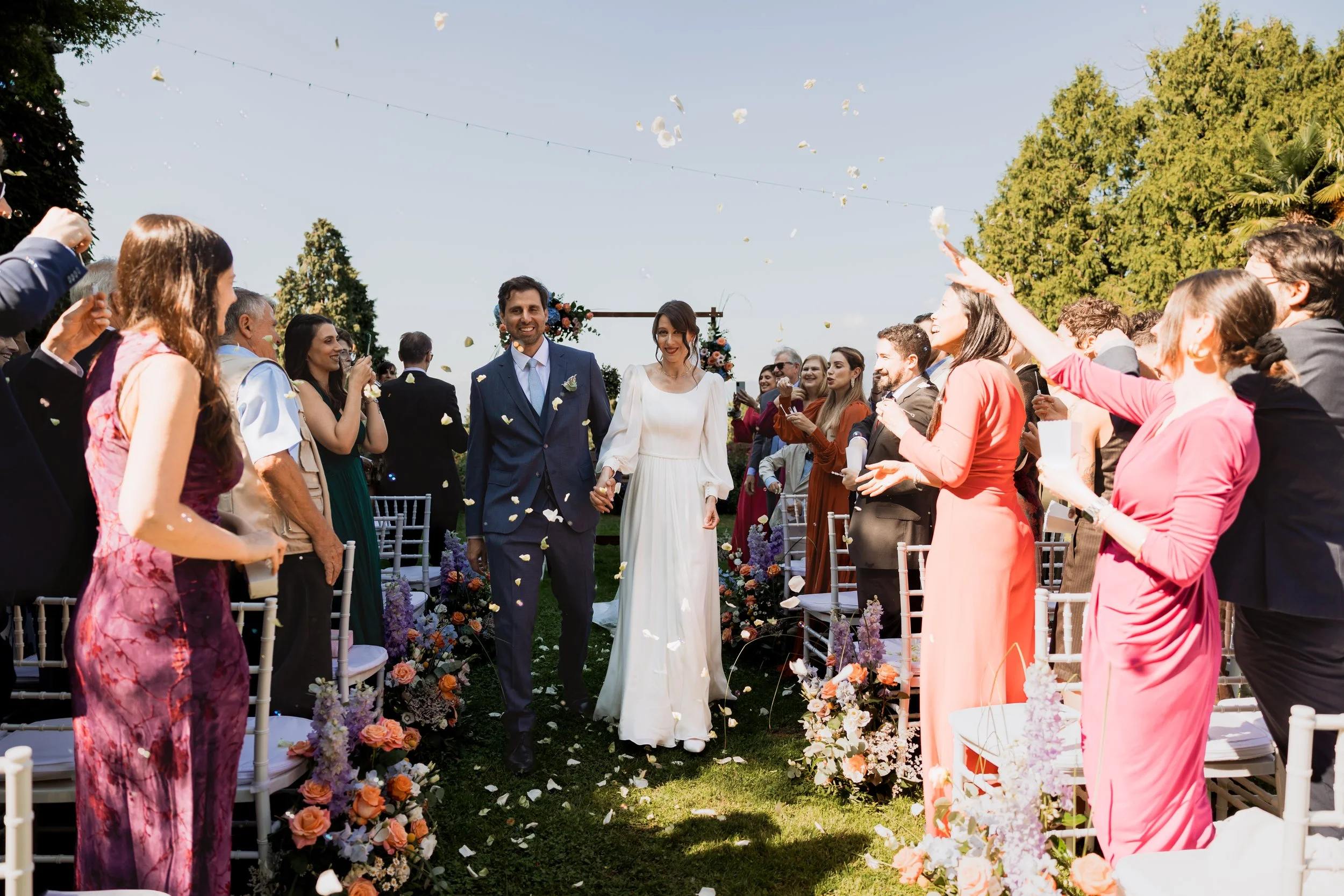 Sposi che camminano tra amici e parenti durante il matrimonio all'aperto con decorazioni floreali e petali di fiori che volano nell'aria.