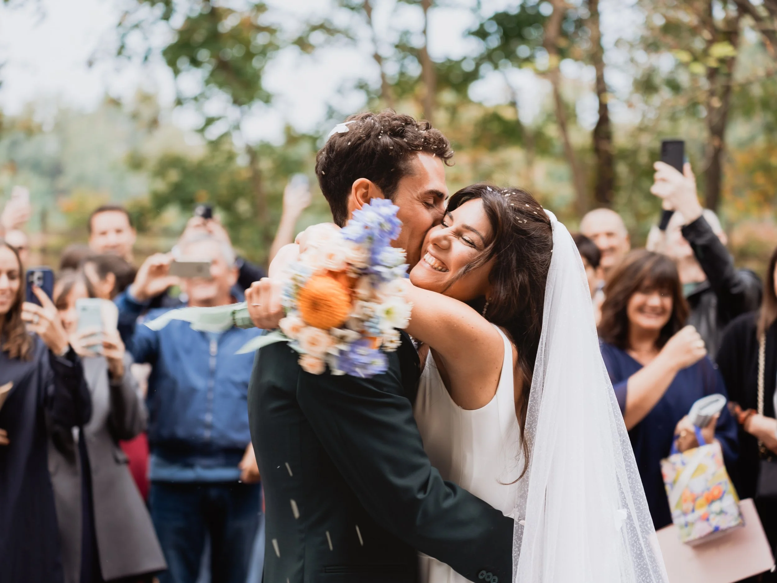 Due persone che si abbracciano e si baciano durante un matrimonio all'aperto, circondati da amici che scattano foto e sorridono.