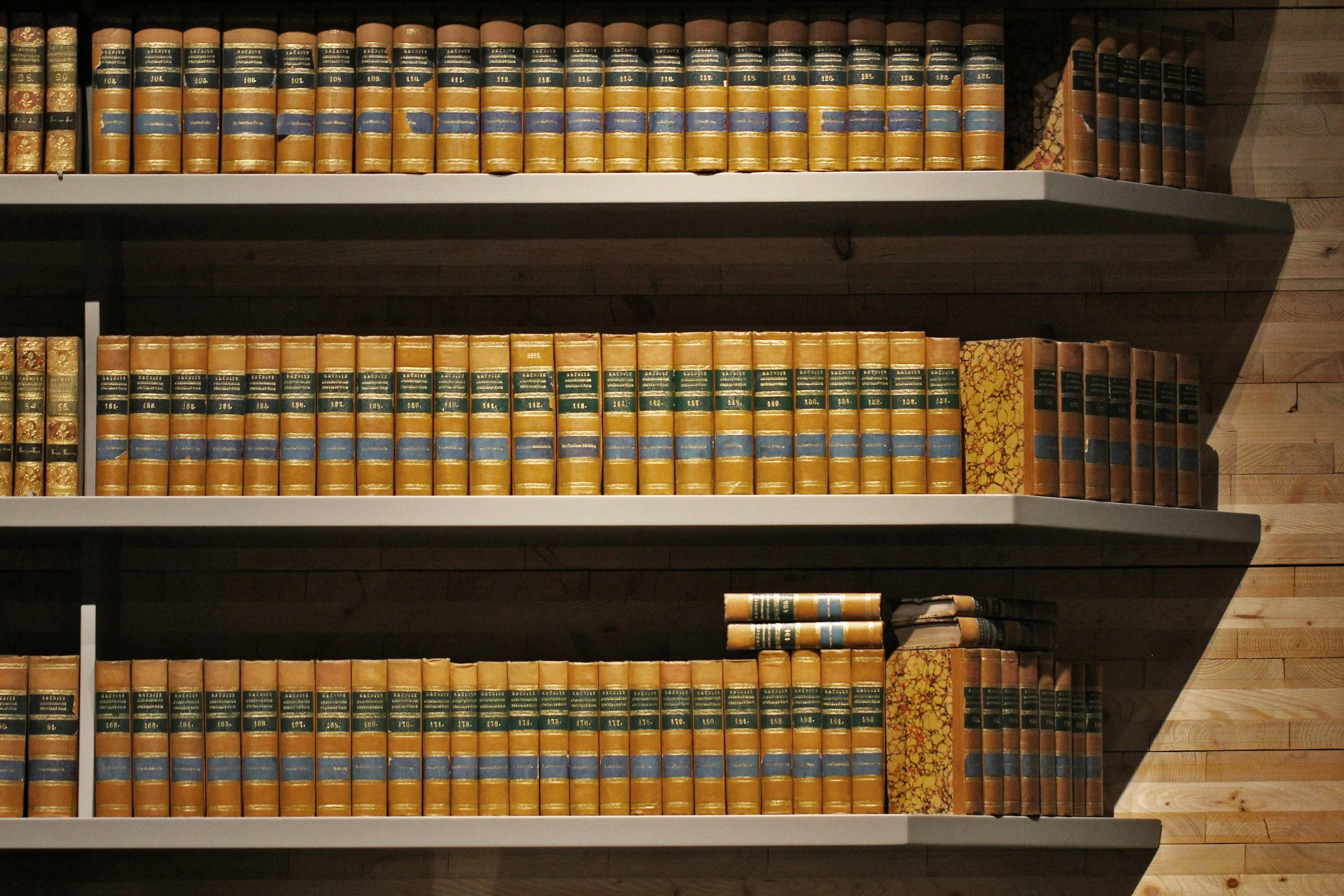 Bookshelf filled with vintage, leather-bound books with gold accents and green labels, arranged on multiple shelves against a wood-paneled wall.